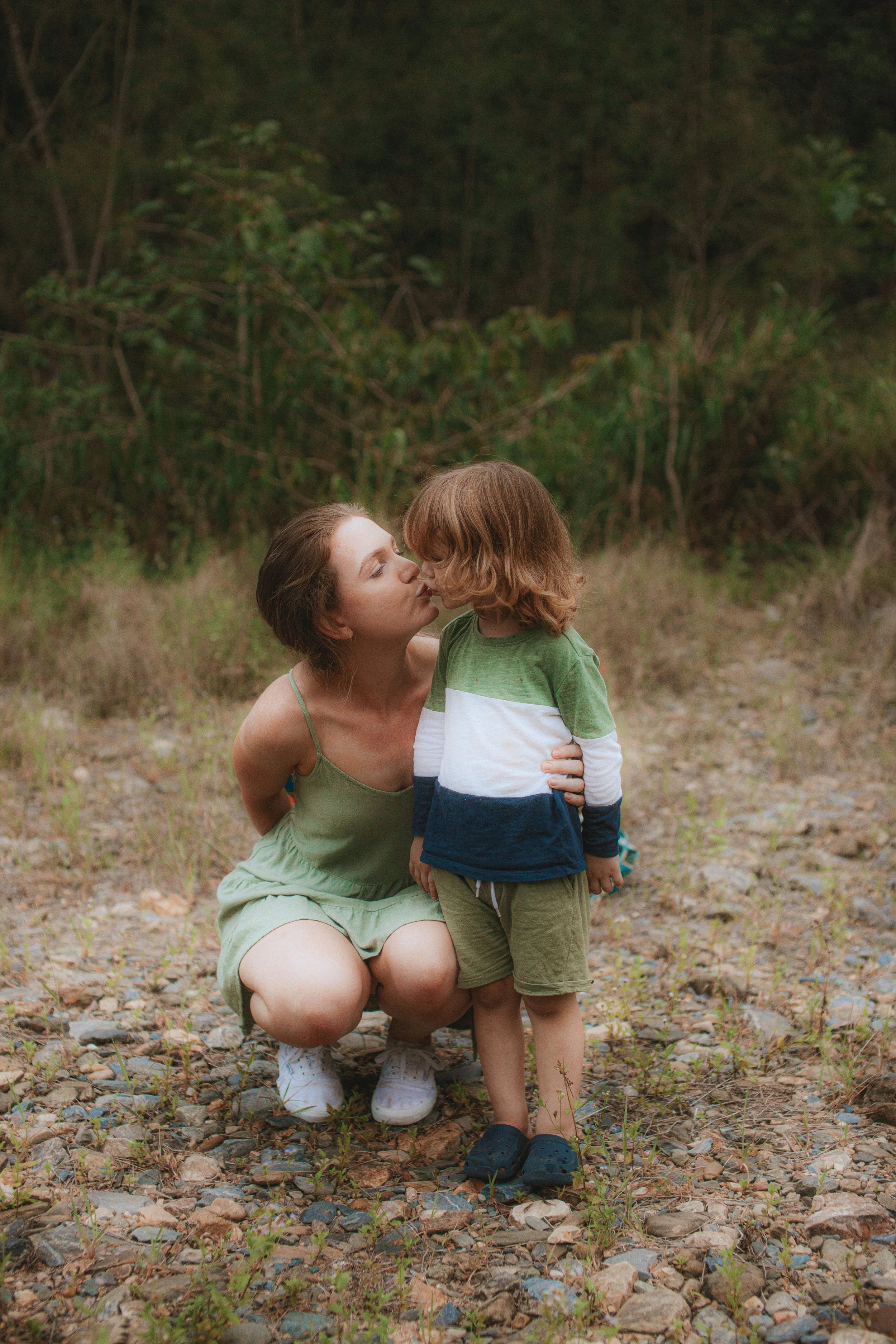 A woman in a green dress crouching down and kissing a young boy in a green and striped shirt on the nose outdoors on a rocky path with trees in the background.