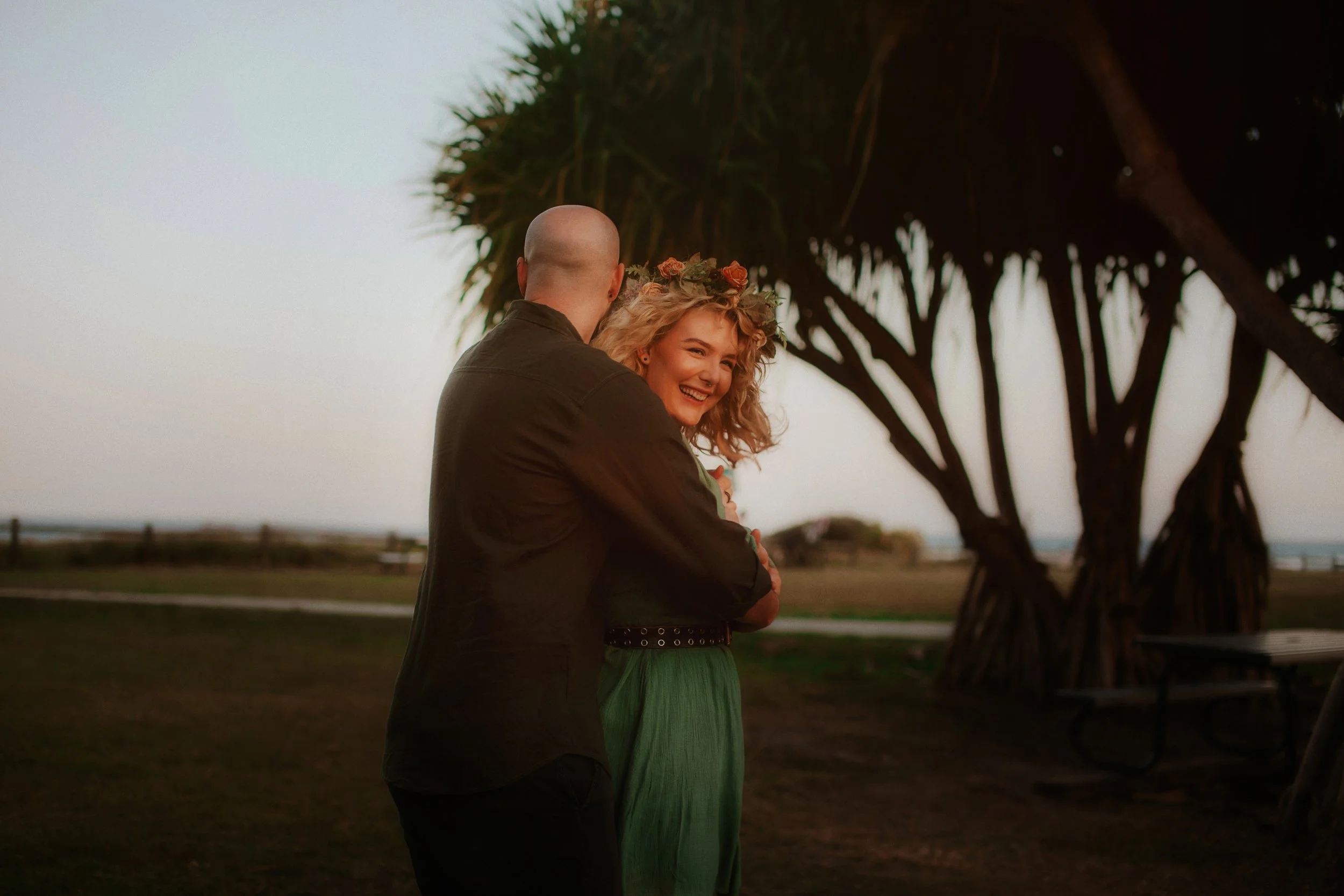 A couple stands outdoors at sunset, with the woman smiling and wearing a flower crown, while the man faces away from the camera.