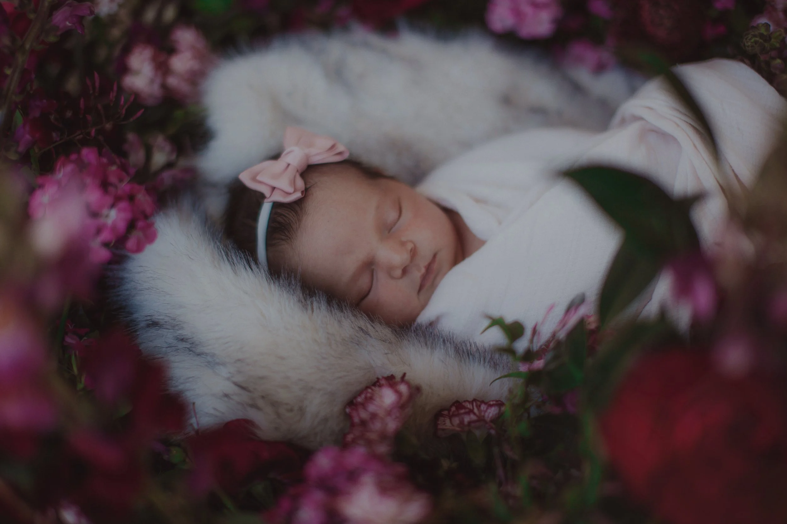 A sleeping newborn baby girl with a pink bow headband, nestled on a fluffy blanket surrounded by pink flowers.