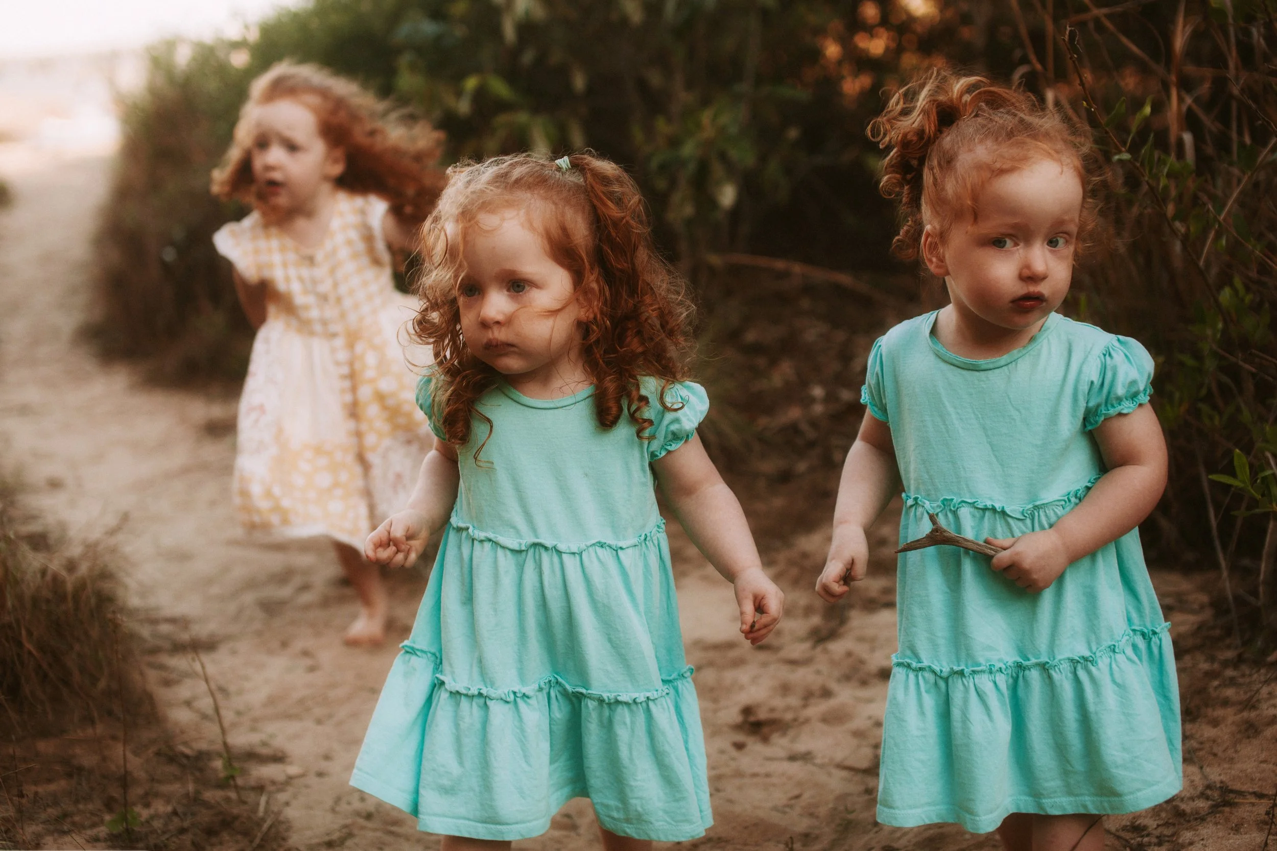 Three young girls with red hair walking outdoors on a dirt path, two dressed in mint green dresses and one in a yellow and white polka dot dress, surrounded by greenery.