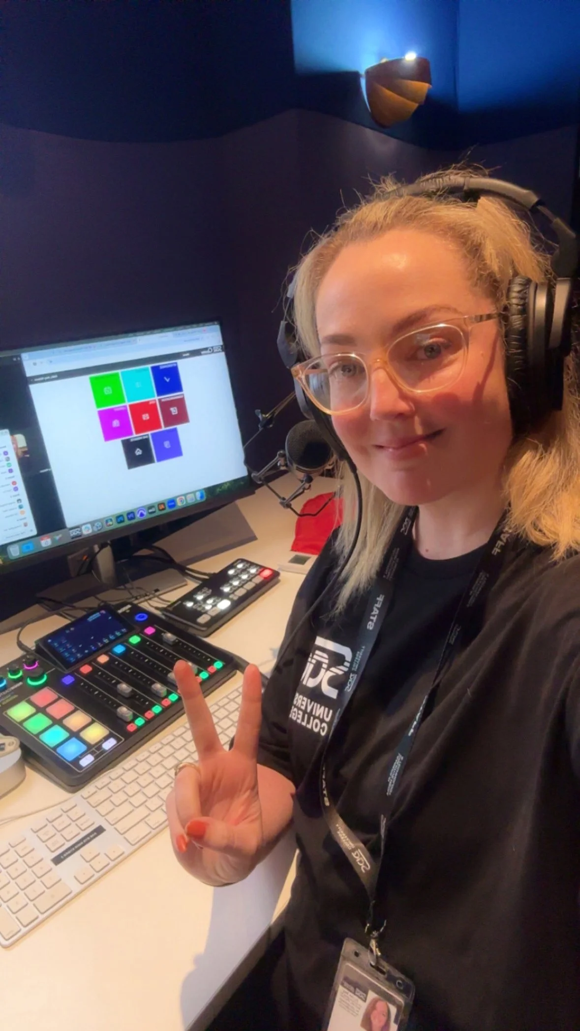 A woman wearing glasses, headphones, and a black shirt with a lanyard is taking a selfie at a workspace with music production equipment. She is making a peace sign with her right hand. There is a computer monitor displaying colorful squares, a keyboard, and various audio devices on the desk.