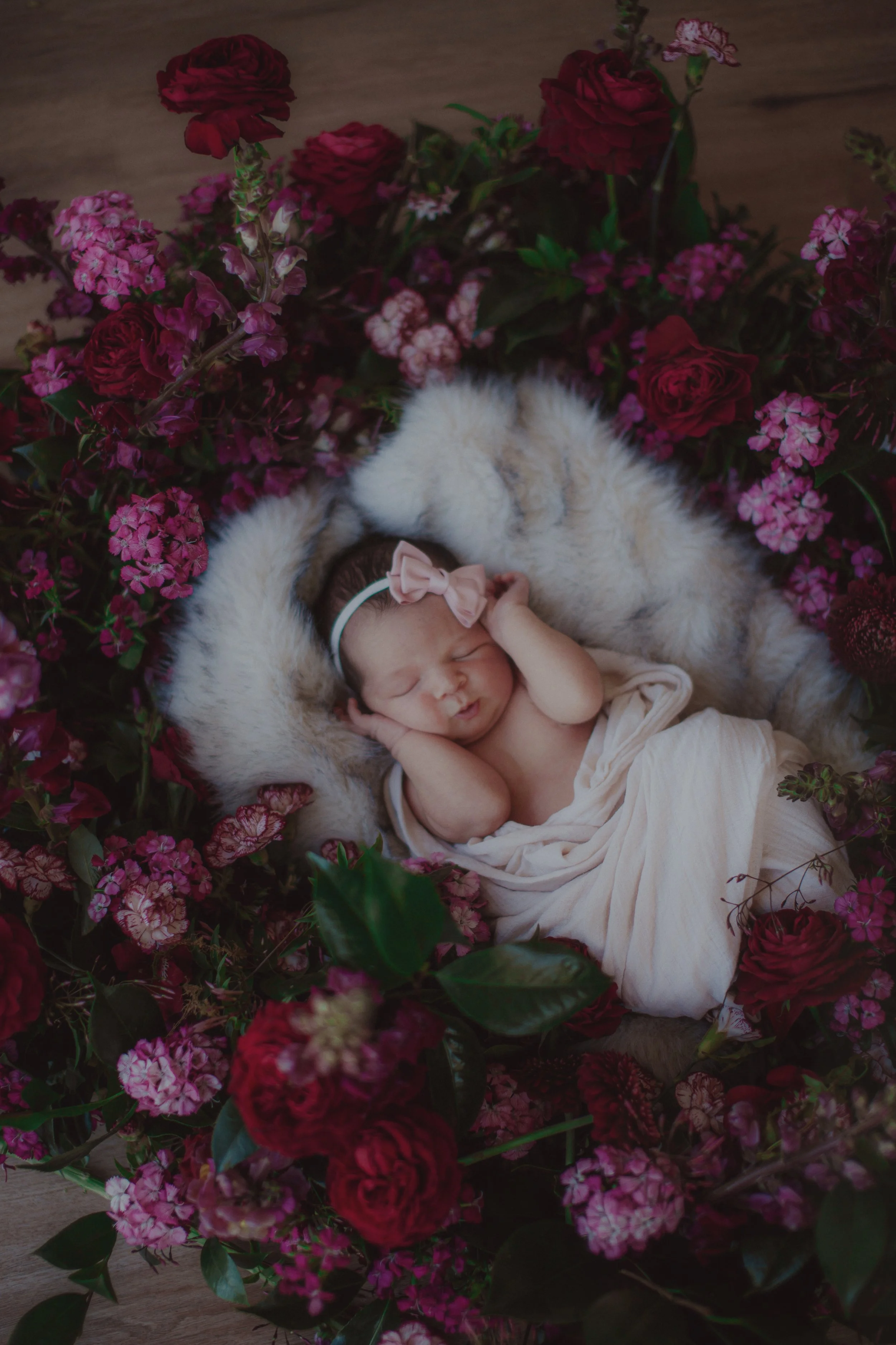 A newborn baby sleeping on a soft fur blanket surrounded by pink and red flowers.