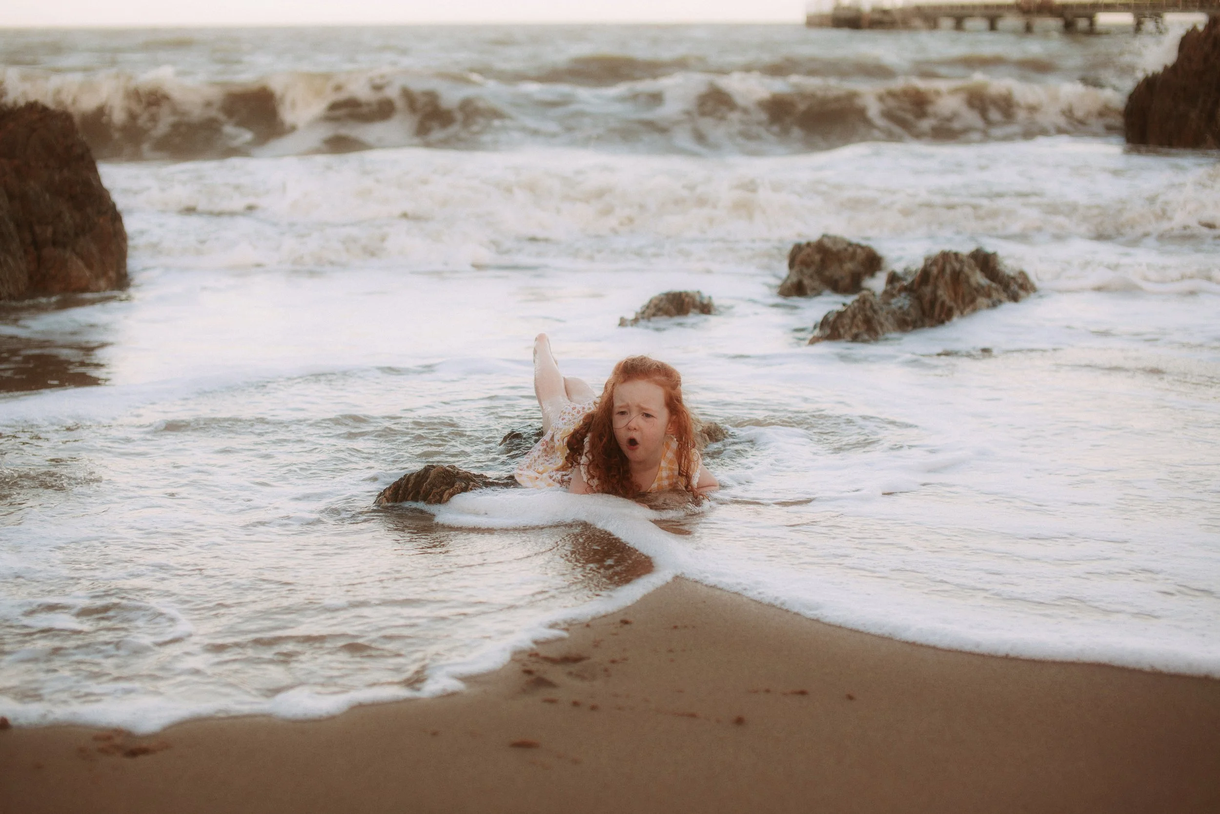 A young girl with red hair lying on her stomach on the beach, partially in the water, with rocks and waves in the background, making an surprised or shocked facial expression.