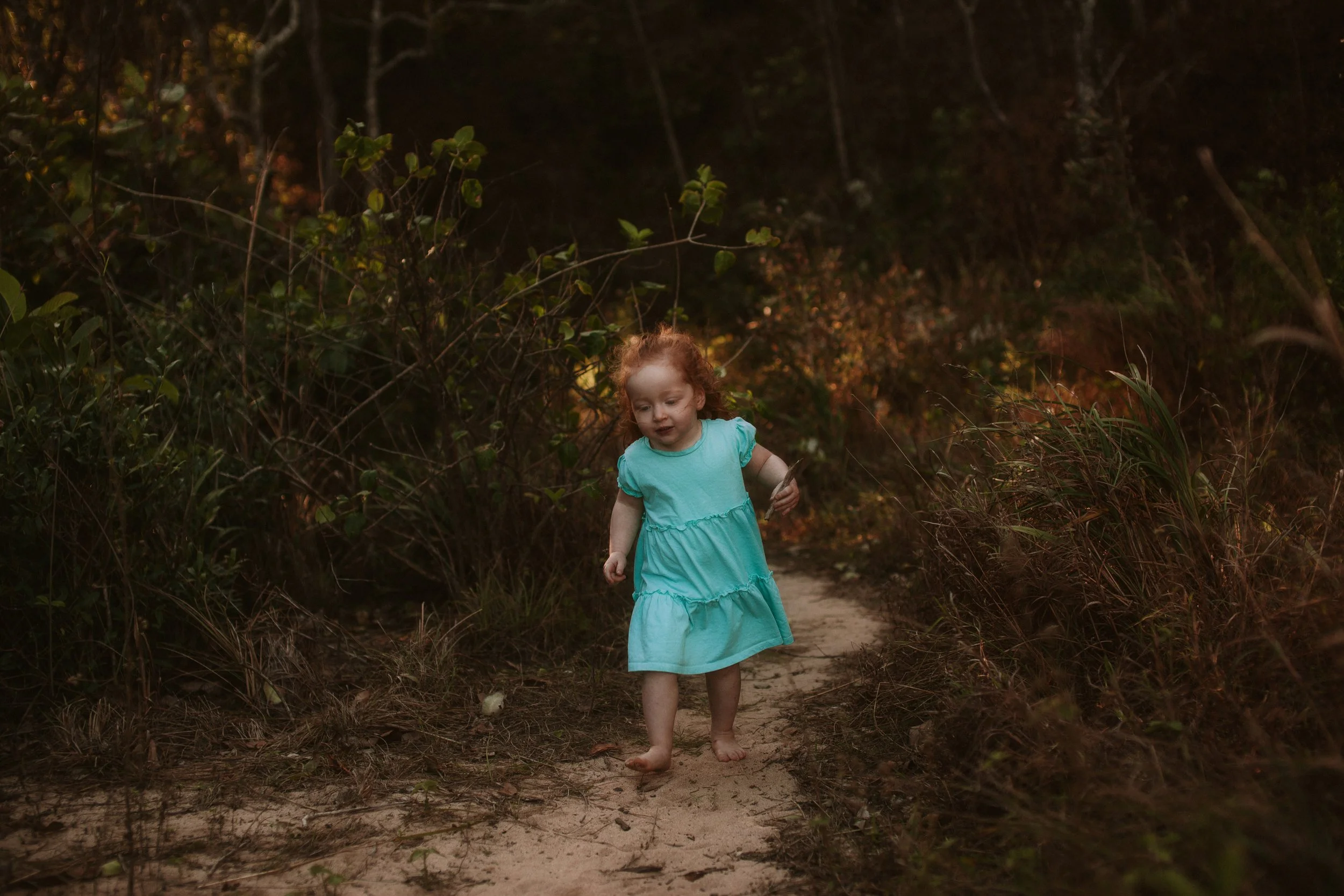 A young girl with red hair wearing a light blue dress walking barefoot on a dirt trail through a wooded area during dusk.