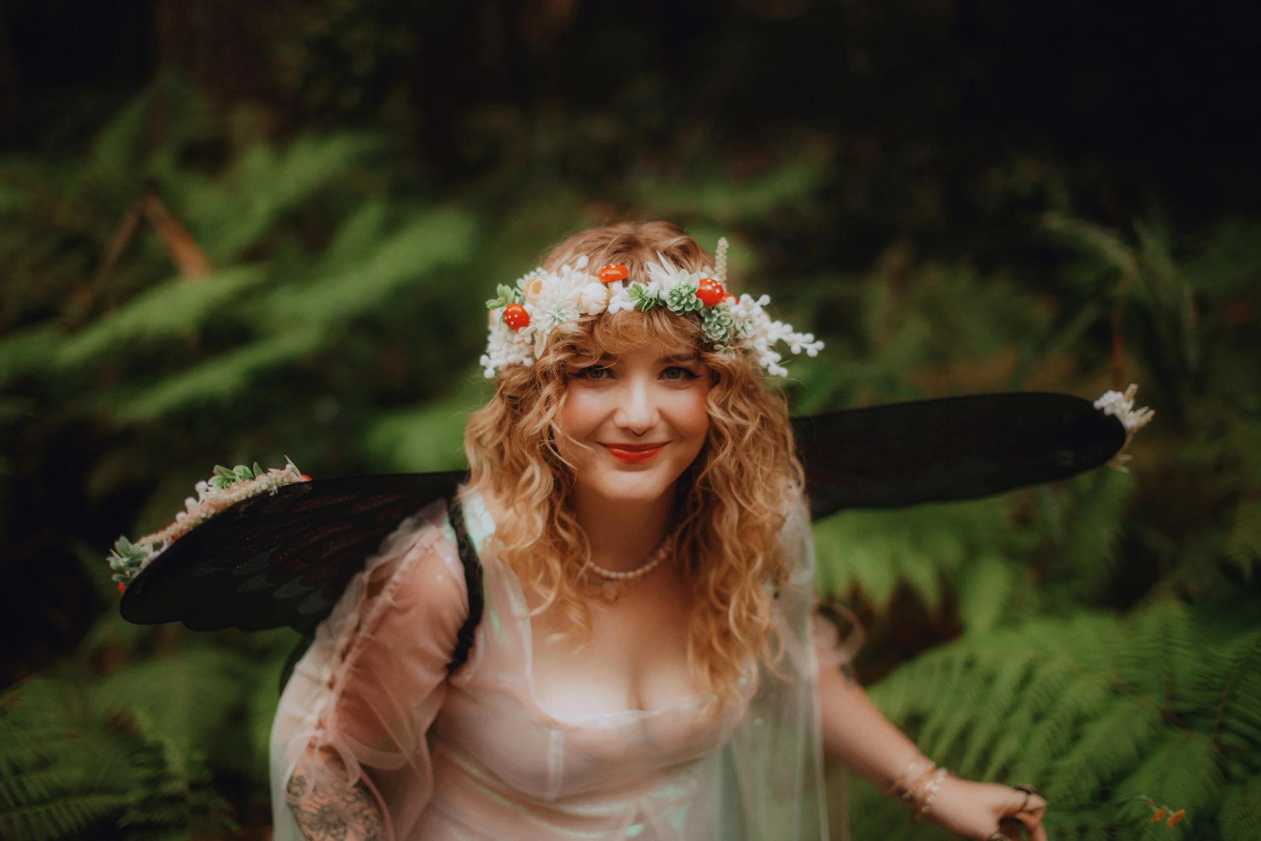 A woman with curly blonde hair, wearing a flower crown with white, red, and green flowers, smiling in a lush green forest. She has fairy wings and a sheer white dress, accessorized with a pearl necklace and bracelet.