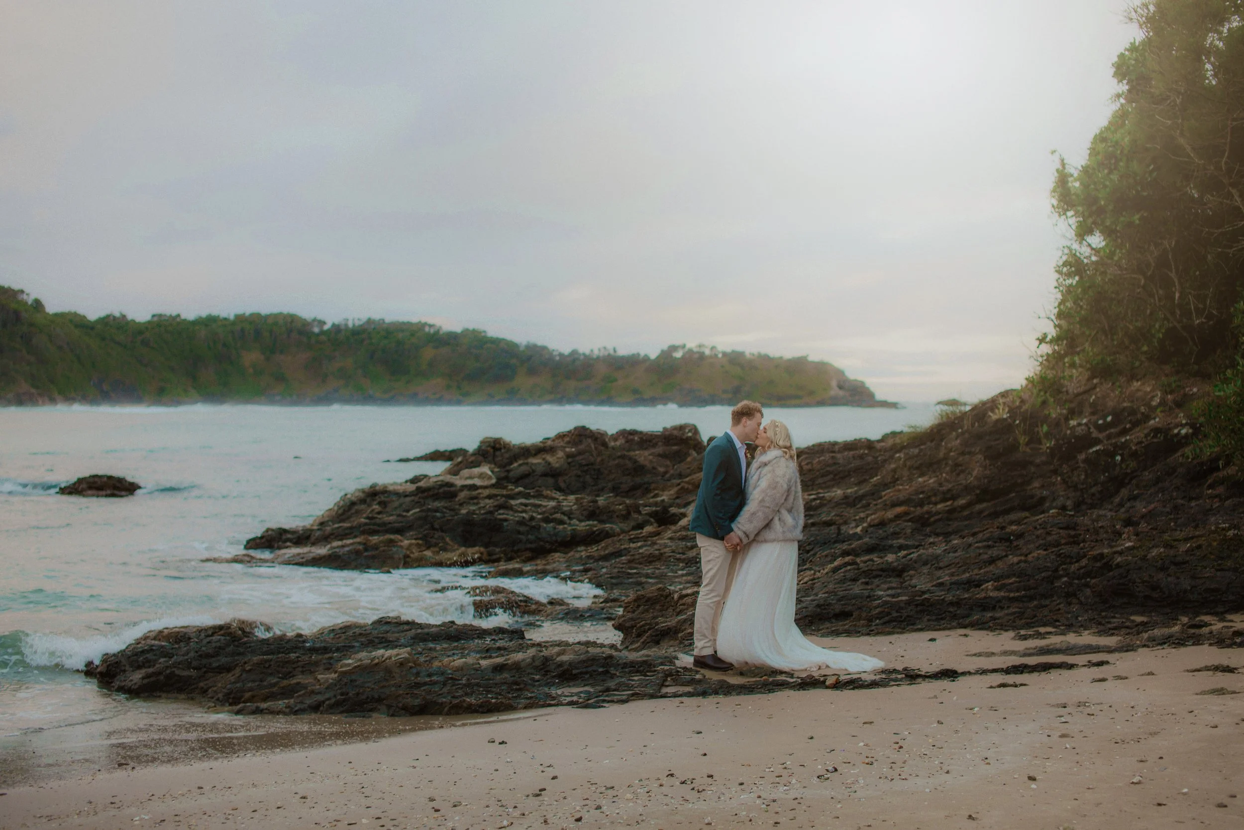 A couple in wedding attire sharing a kiss on a rocky beach during daytime with cloudy skies.
