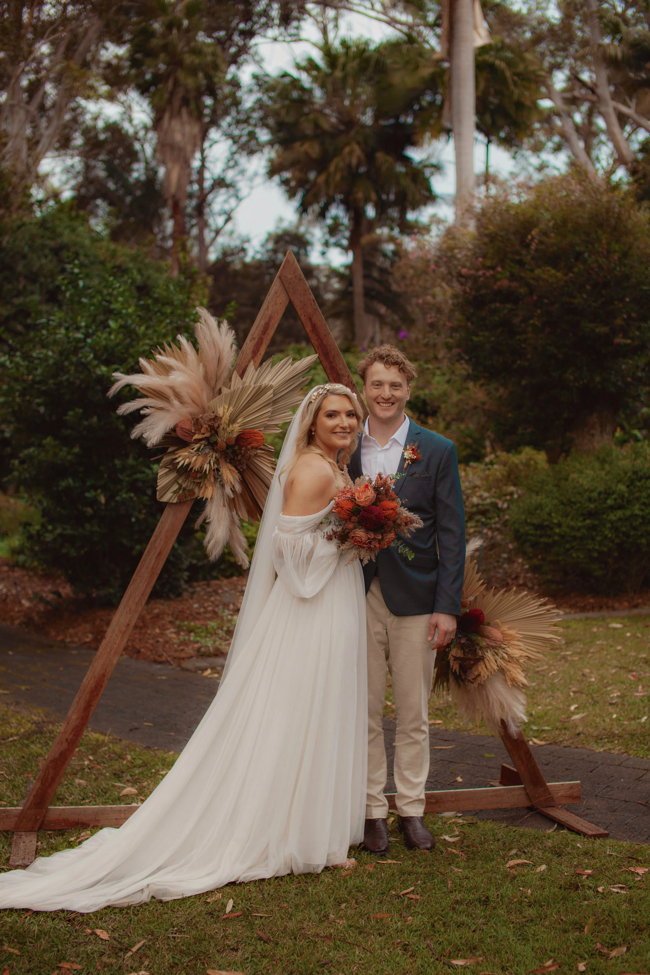 A bride and groom smiling, standing close together outdoors in front of a decorative wooden triangle with dried floral arrangements. The bride wears a white off-the-shoulder wedding gown and veil, and holds a bouquet of dried flowers. The groom wears