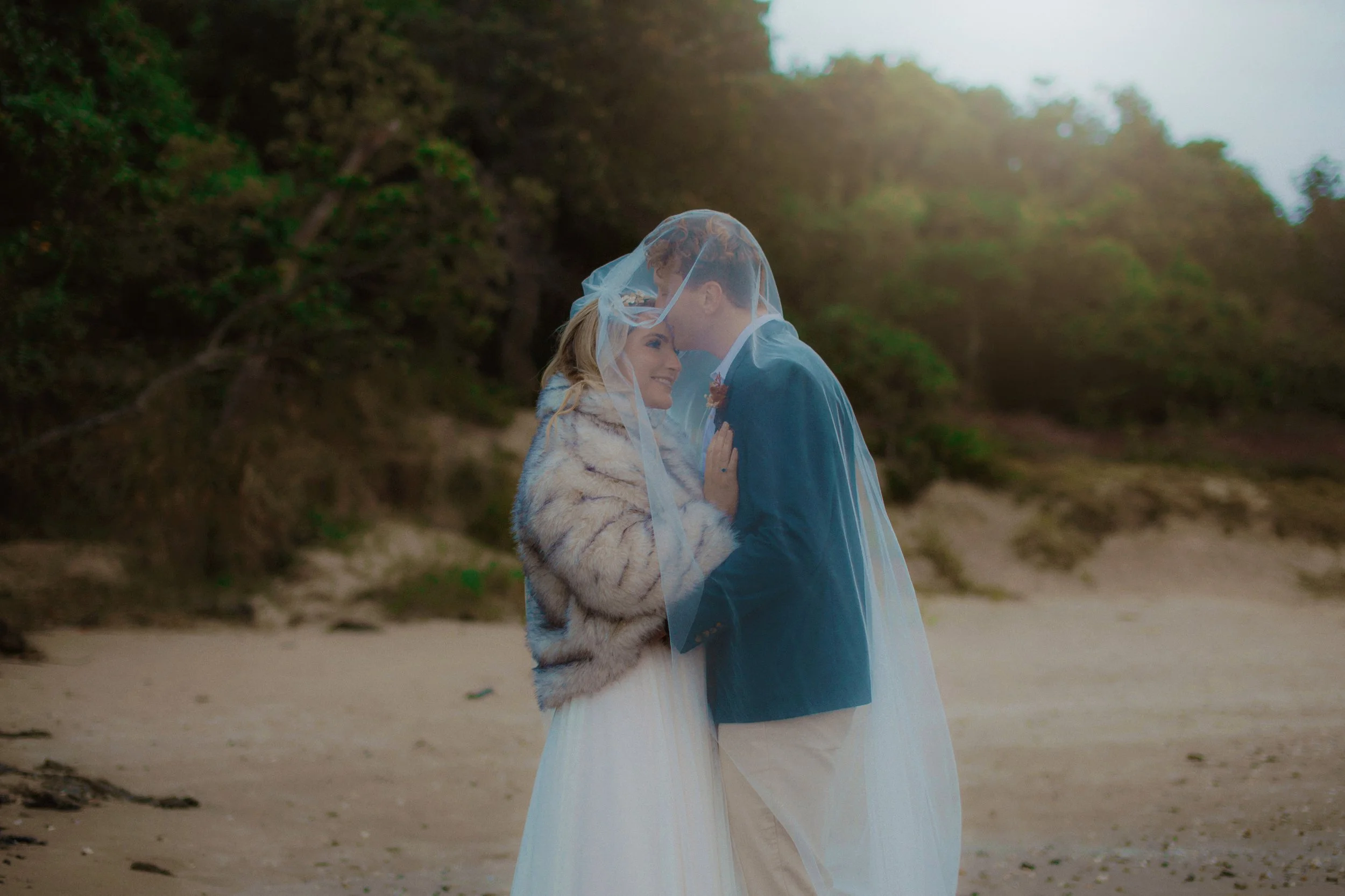 A couple on a beach, with the woman wearing a fur coat and the man in a blazer, sharing an intimate moment under a wedding veil.