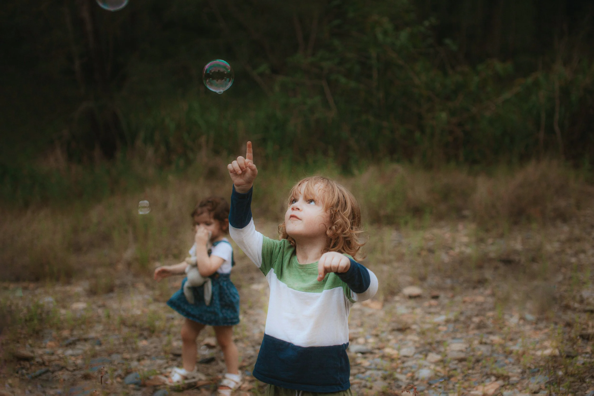 A young boy with curly red hair and a green, white, and navy blue shirt reaching up to touch a floating soap bubble in an outdoor setting with trees and rocks.