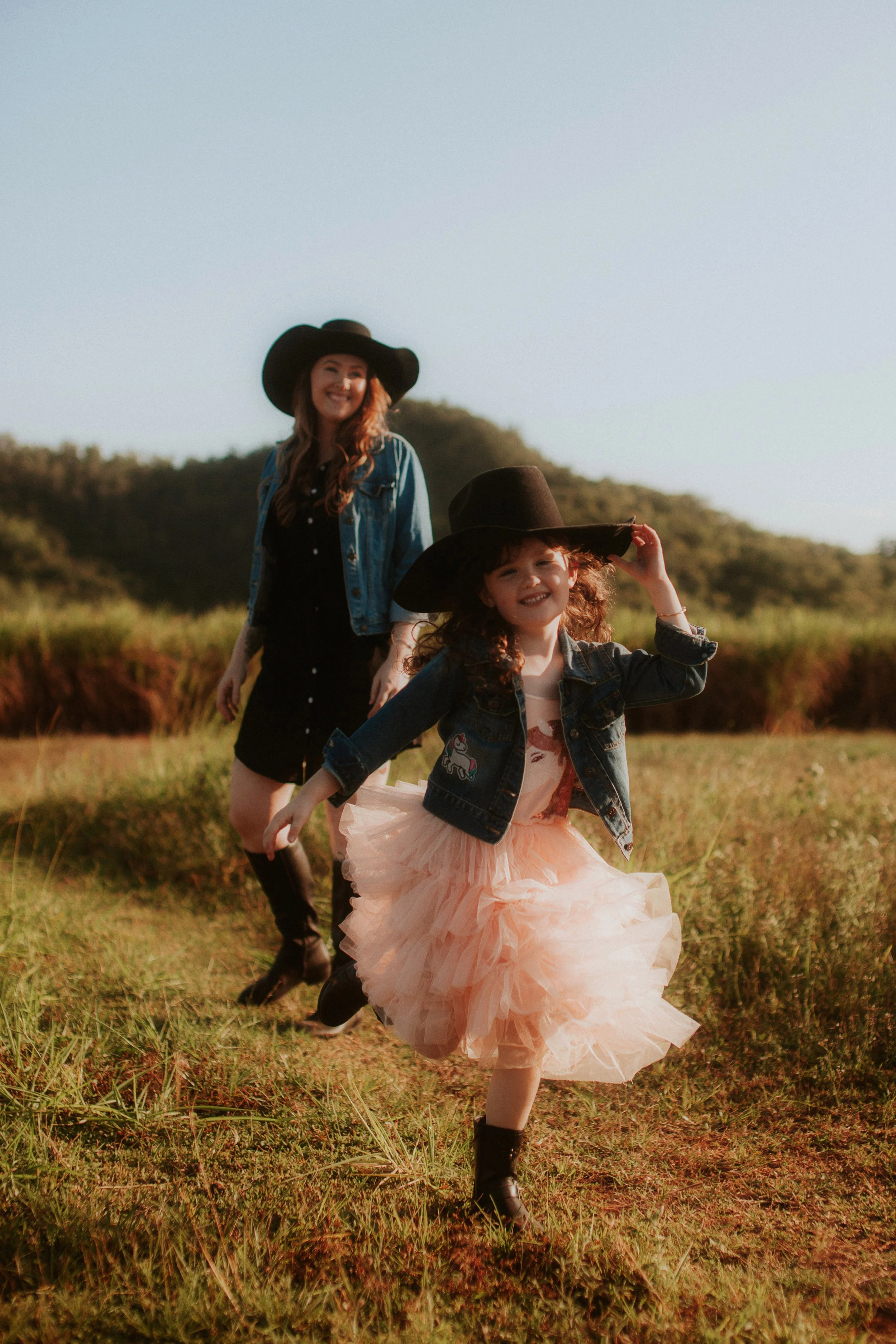 Two women, one young girl and one adult woman, wearing wide-brimmed black hats and denim jackets, running through a grassy field during golden hour. The girl is wearing a peach tutu, black boots, and a denim jacket with patches, smiling and holding h