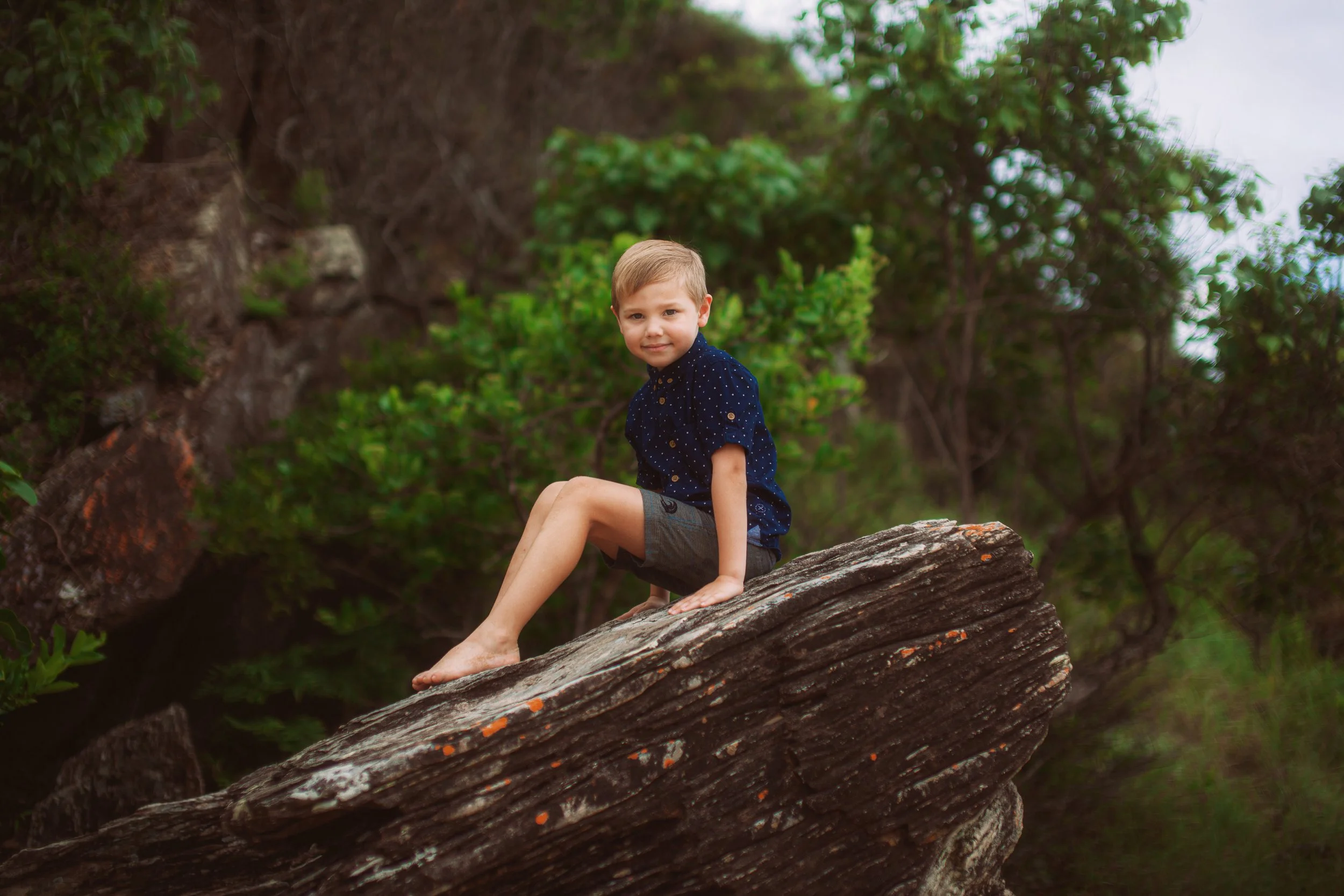 A young boy with blond hair sitting on a large, textured log outdoors surrounded by green trees and foliage.
