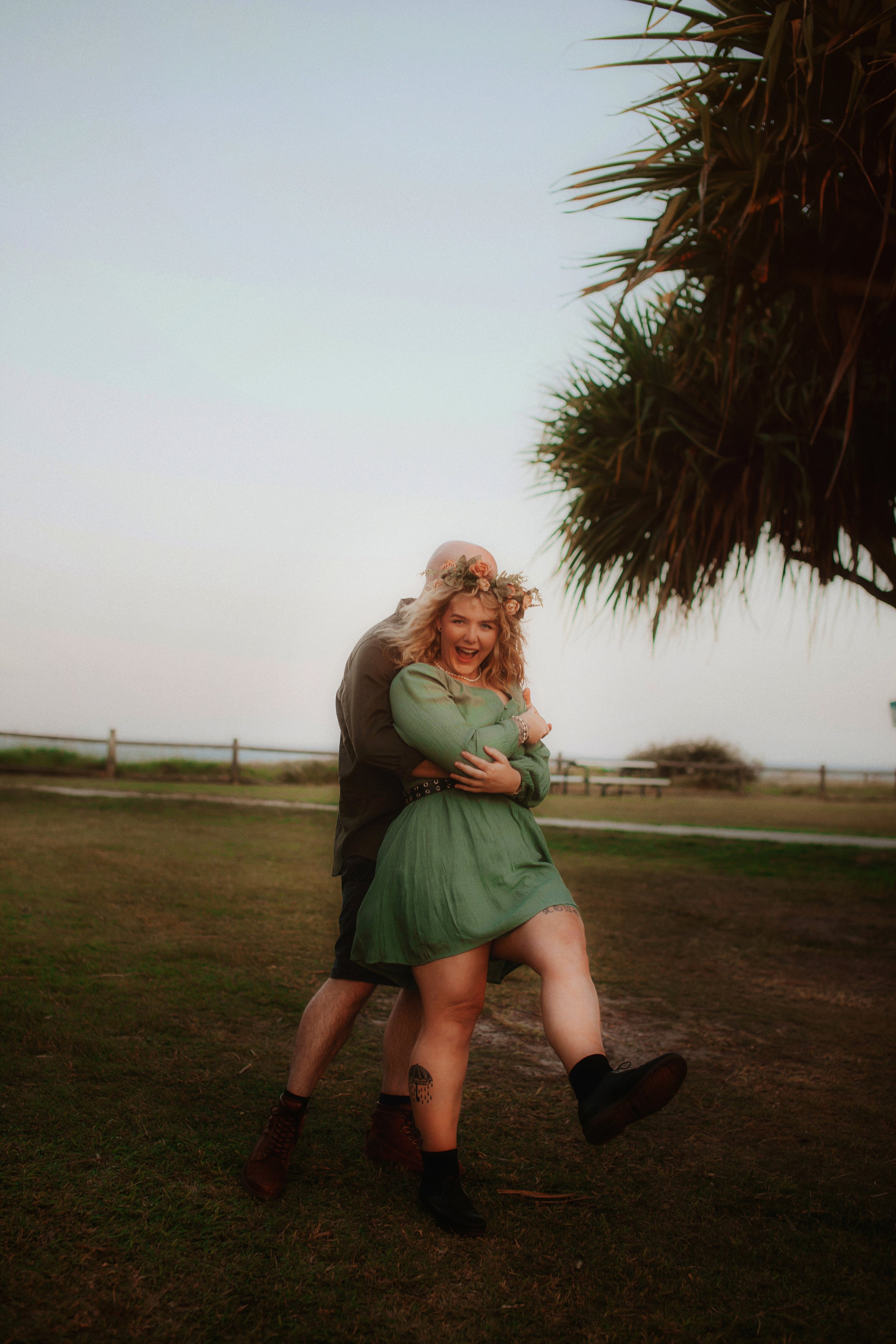 A woman with curly blonde hair wearing a green dress and a floral headband is smiling and laughing as she is lifted off the ground by a man wearing dark clothing. They are outdoors on a grassy area with a tree in the foreground and a fence in the bac