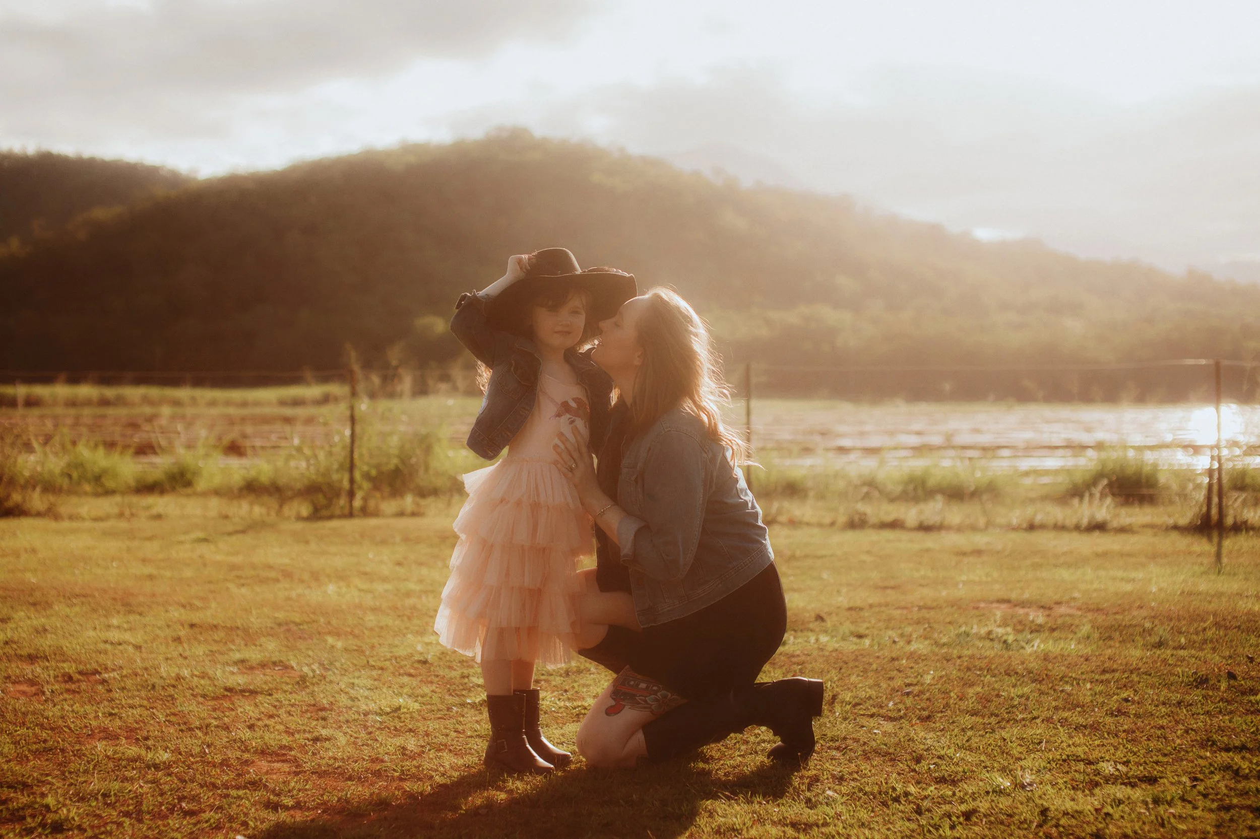 A woman kneeling on the grass and kissing a young girl who is holding a hat on her head. The girl is wearing a pink dress and black boots, and the woman is wearing a denim jacket and black shorts. They are outdoors during sunset with mountains and wa