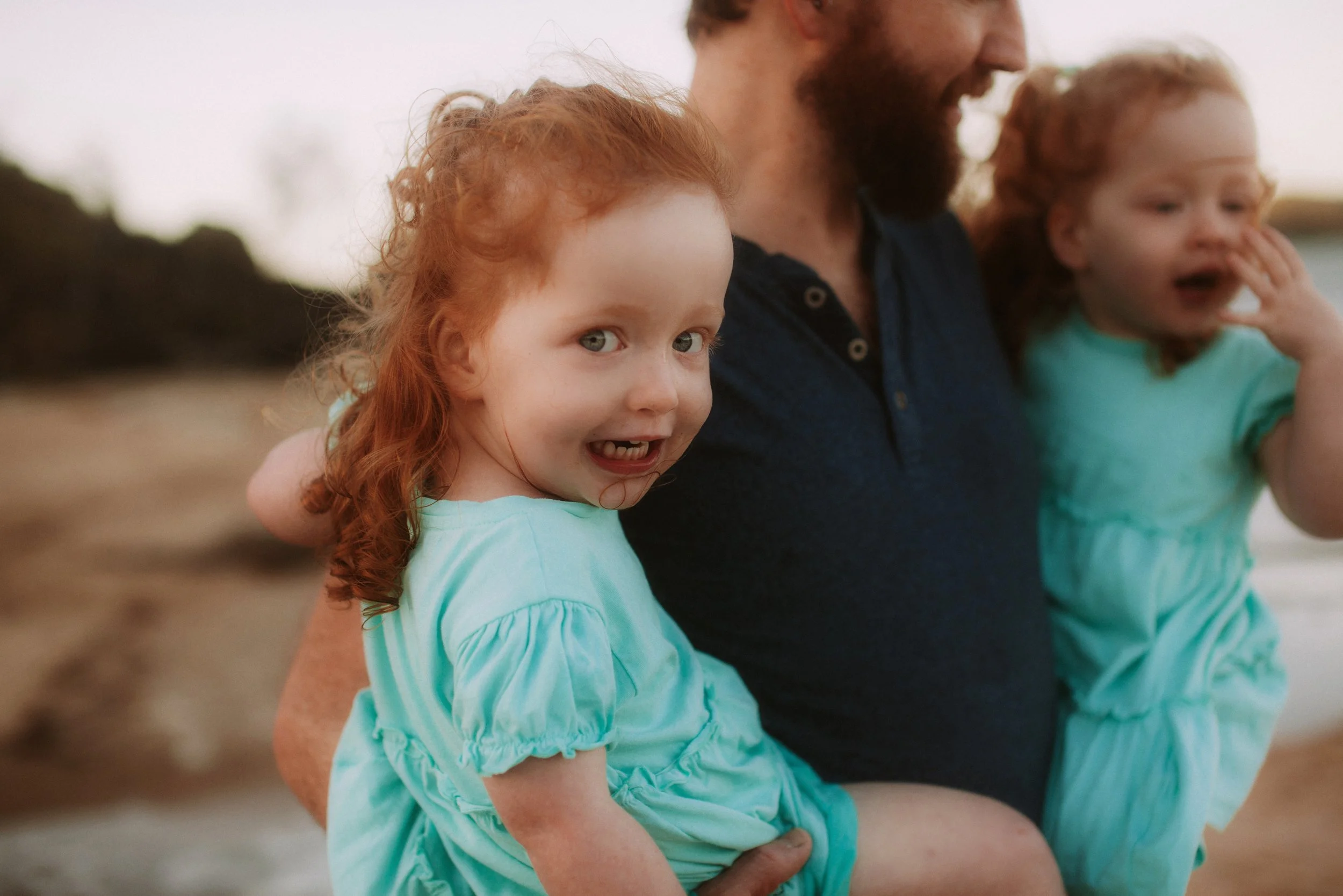 A man with two young red-haired girls with curly hair at the beach during sunset, both wearing light green dresses.