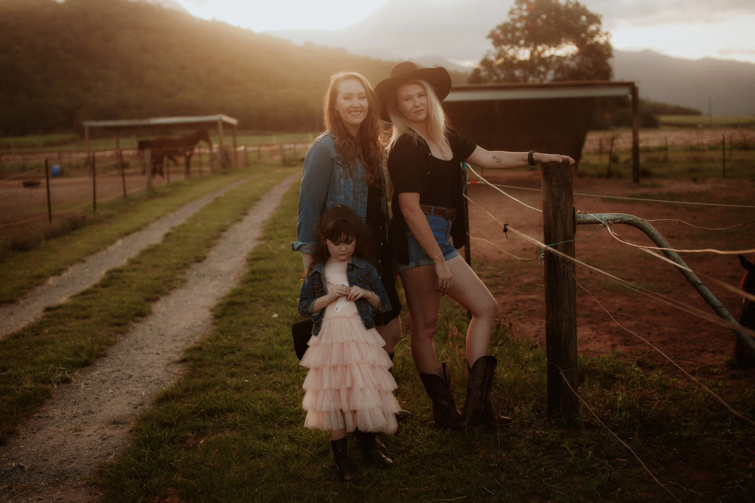 Three women and a young girl standing on a farm at sunset, with a horse in the background and green hills in the distance.