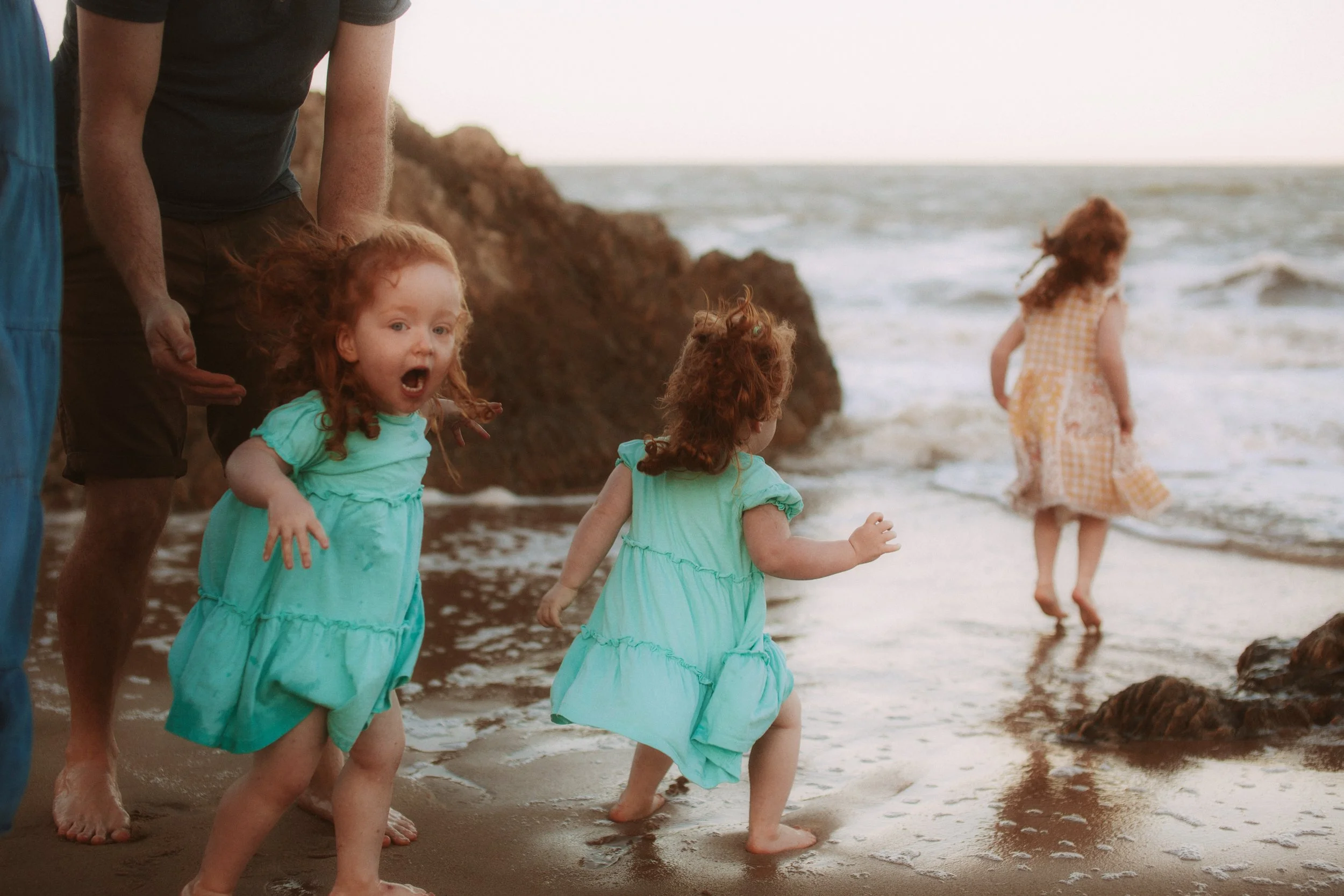 Three young red-haired girls in turquoise dresses playing by the ocean with a man nearby, one girl running towards the water and the other looking surprised with her mouth open.