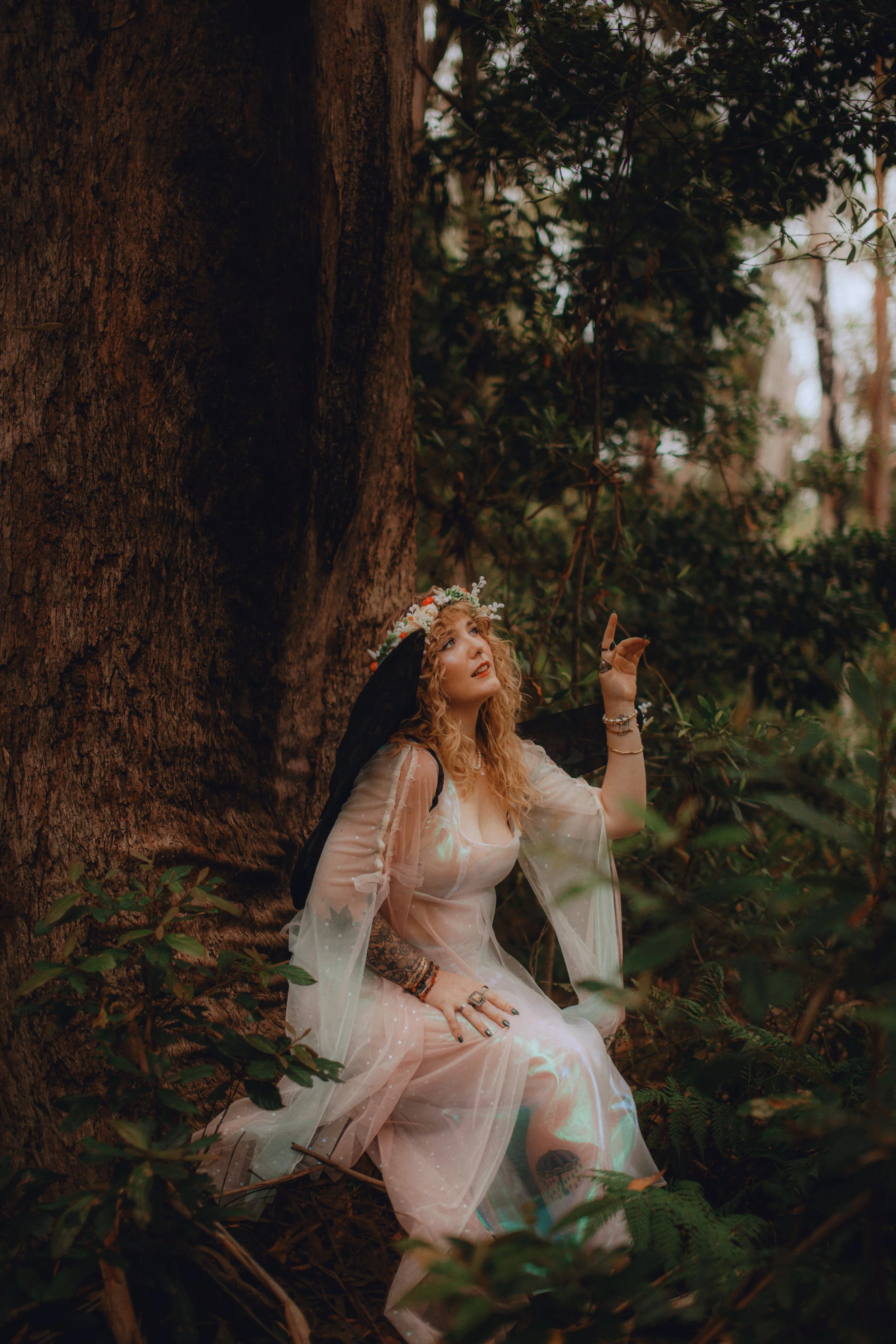 A woman with curly blonde hair wearing a floral crown and a sheer, flowing dress sits on the forest floor near a large tree, looking upwards and pointing with her right hand, surrounded by greenery and branches.