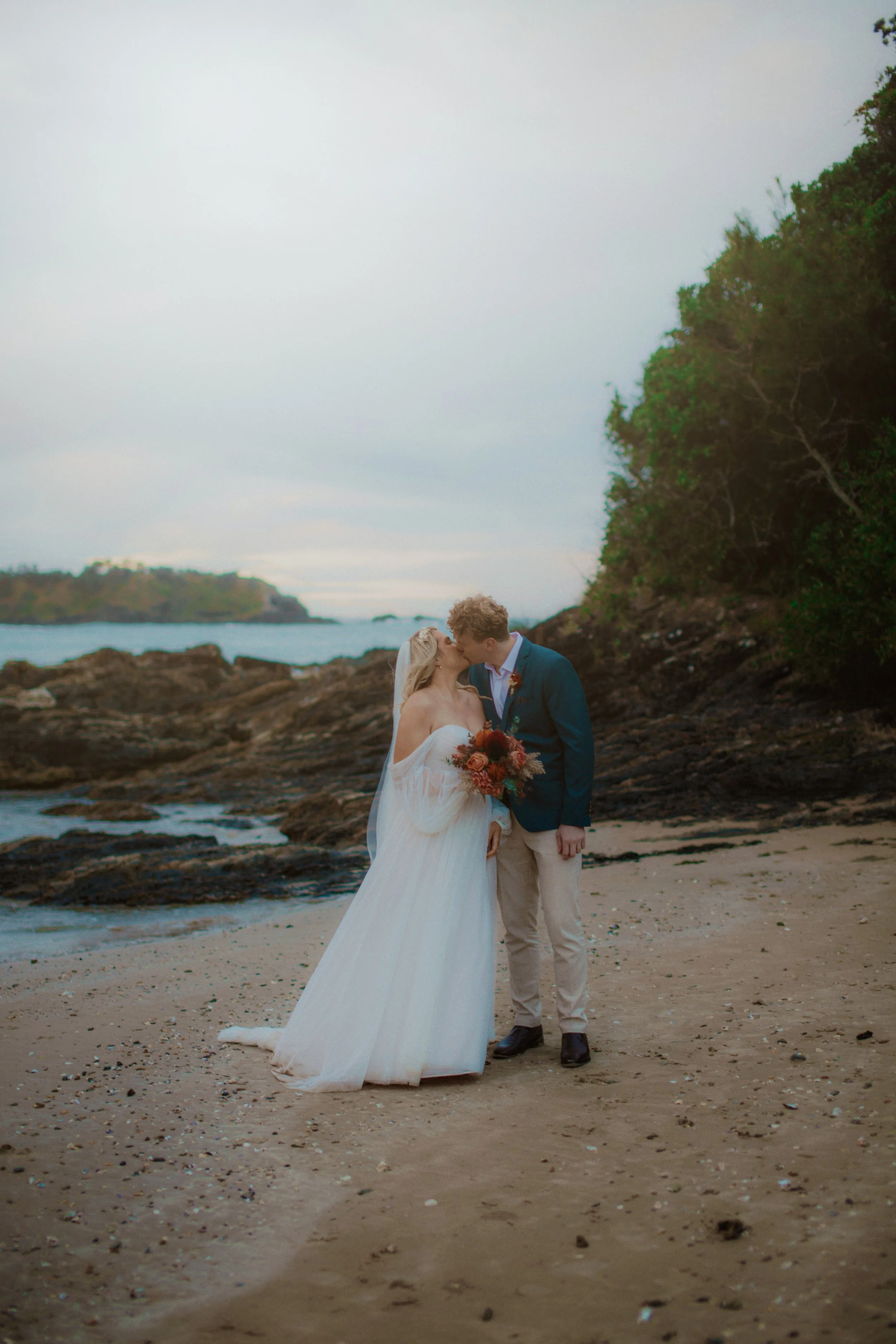 A bride and groom share a kiss on a beach during their wedding, with one holding a bouquet of flowers and the ocean, rocks, and greenery in the background.