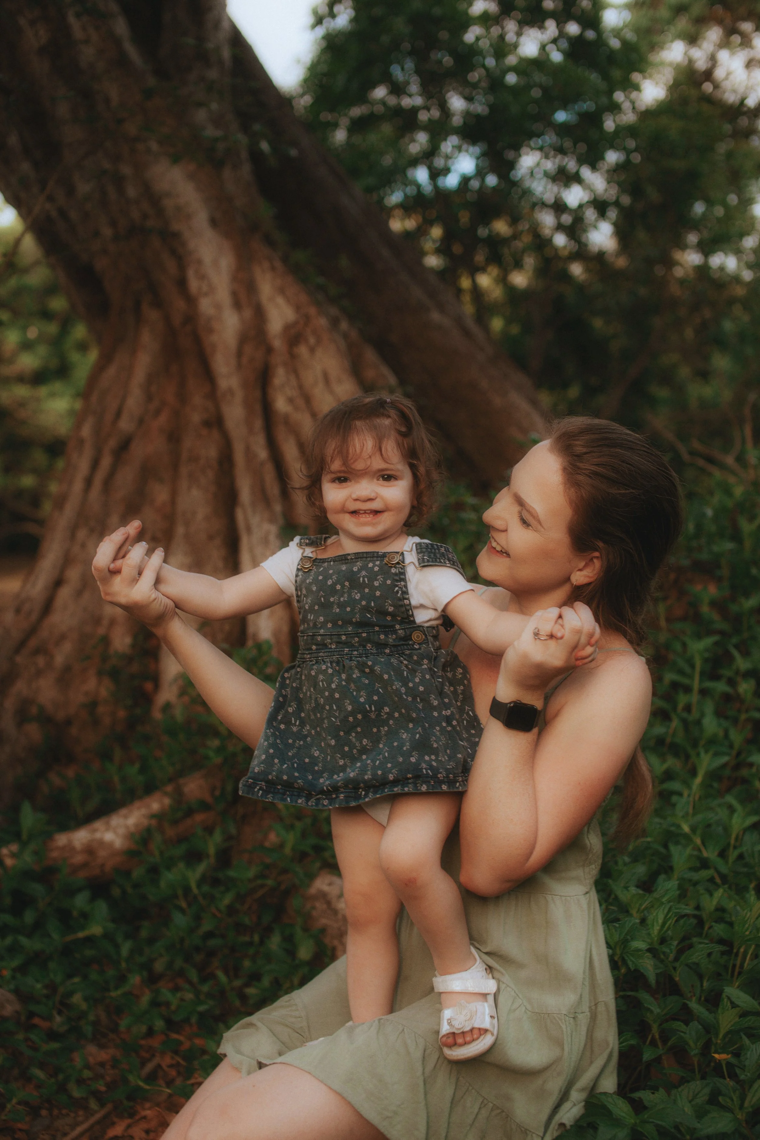 A woman holding a smiling young girl in front of a large tree outdoors.