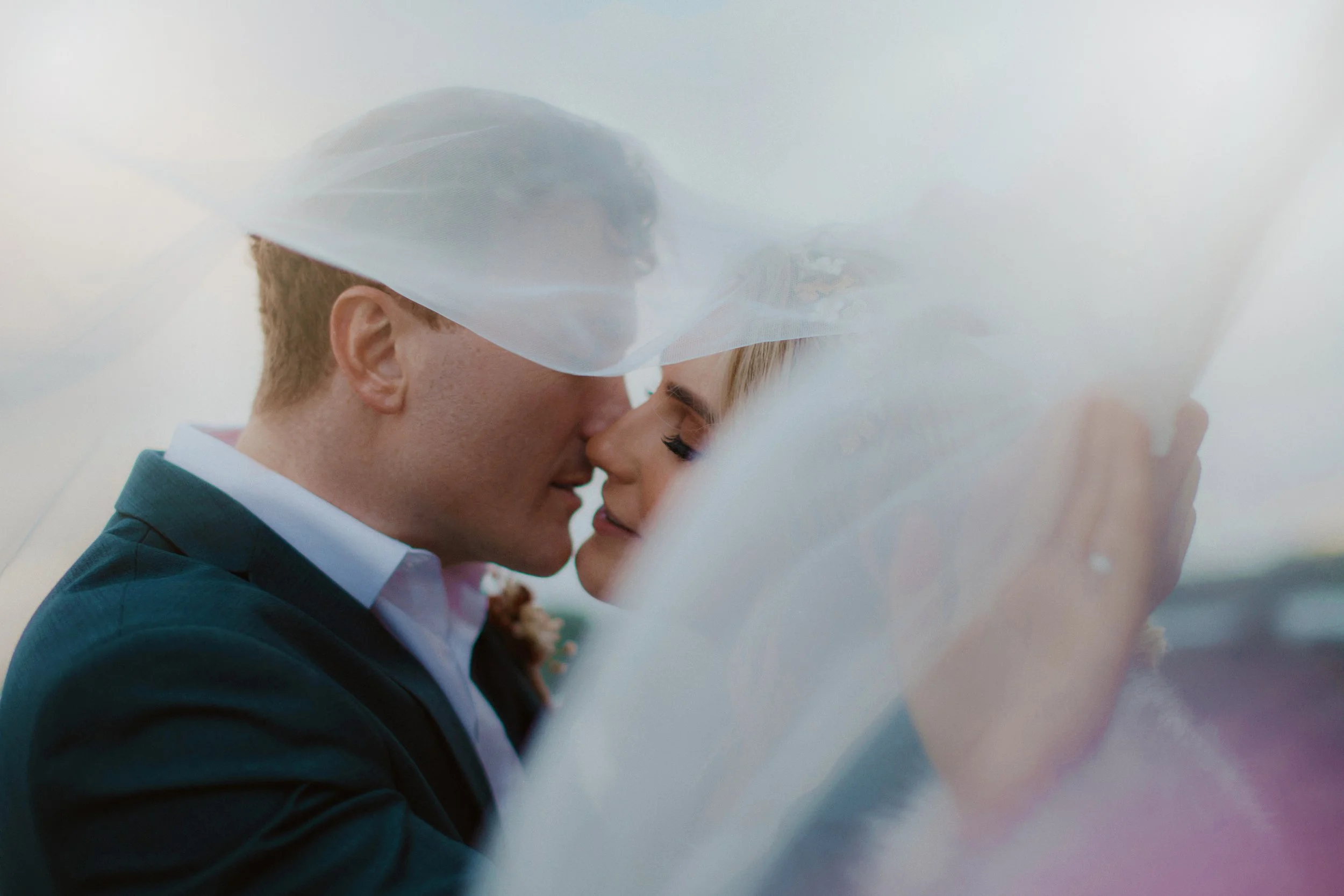 A romantic moment between a bride and groom, their faces close together, under a veil with a soft background.