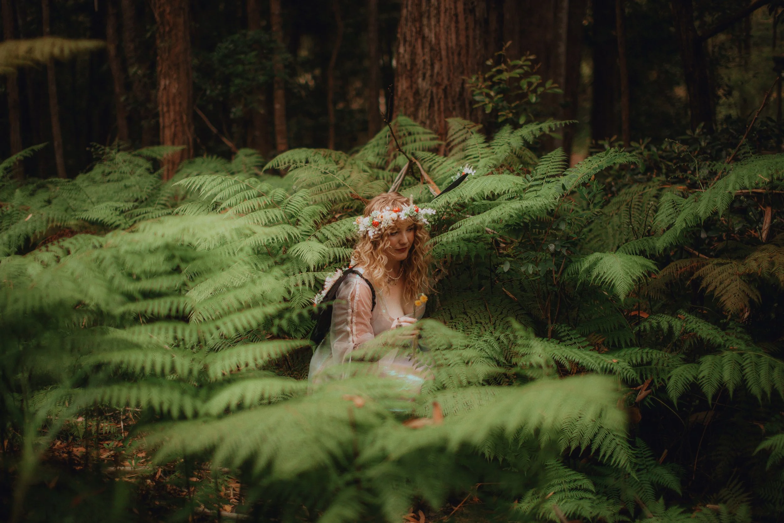 A woman with curly blonde hair wearing a flower crown, a light pink dress, and a backpack, is standing amidst lush green ferns in a forest.
