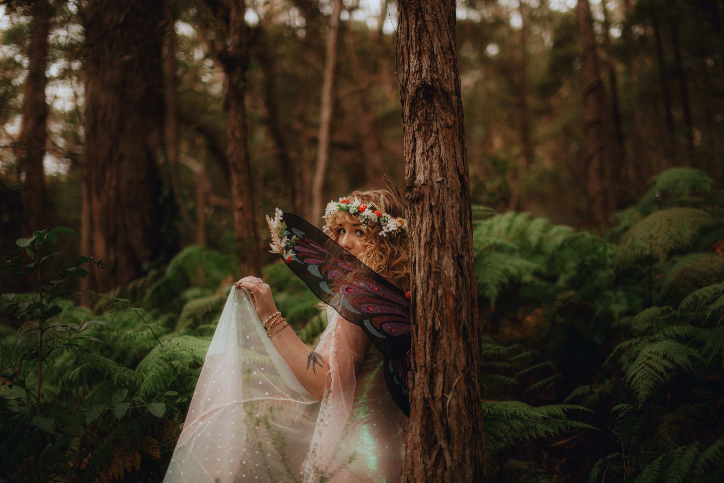 A woman with fairy wings, wearing a flower crown, standing behind a tree in a forest, holding a flowing veil, looking over her shoulder.