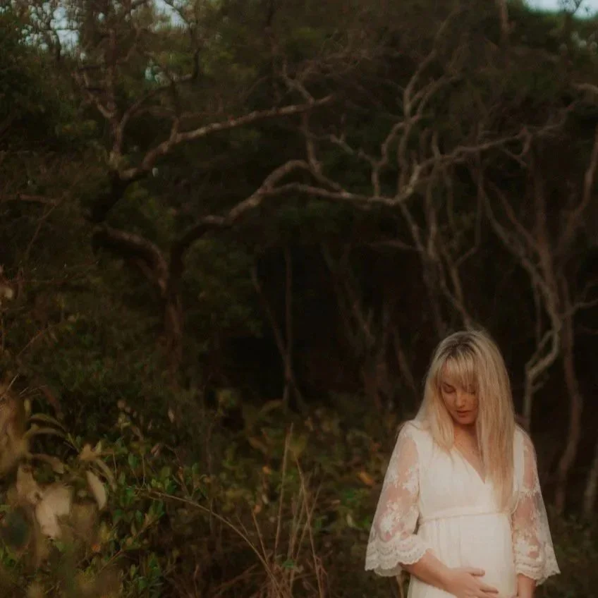 A pregnant woman with blonde hair wearing a white lace dress, standing outdoors in a forested area with twisted tree branches and dense greenery in the background.