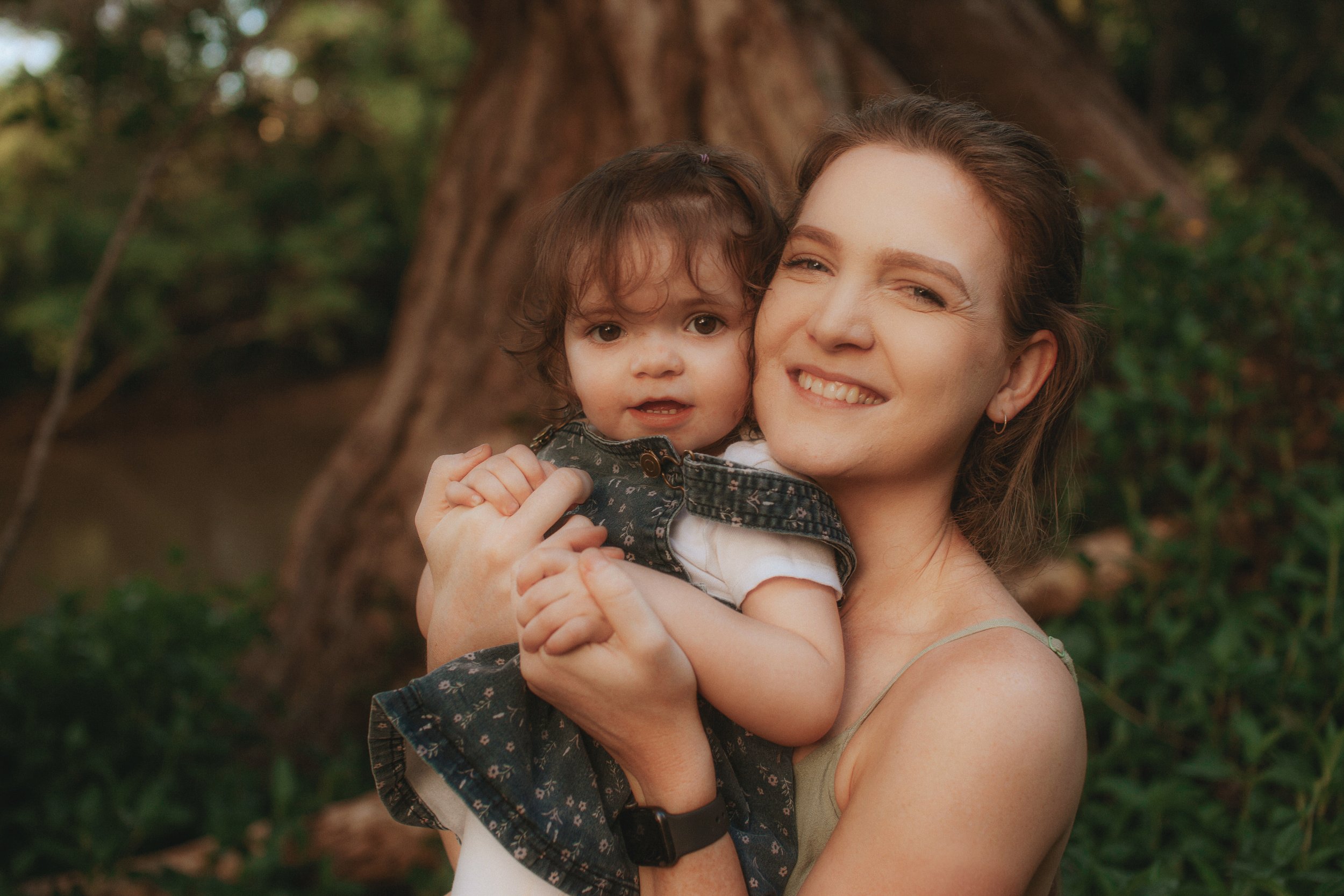A woman holding a young girl outdoors near a tree, both smiling at the camera.