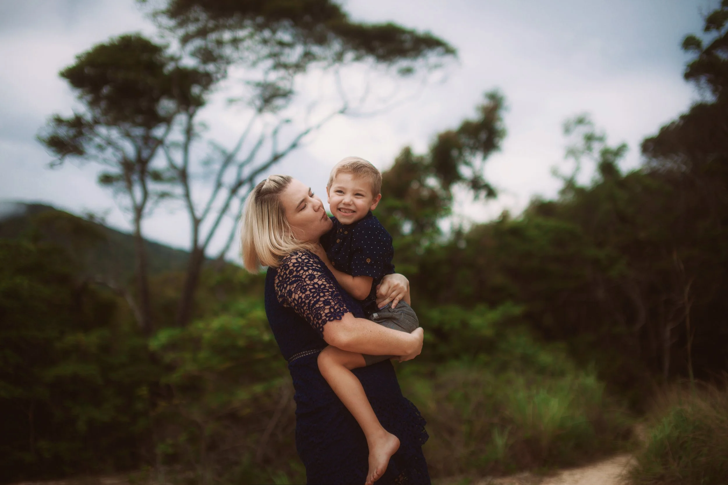 A woman holding a young boy outdoors in a natural setting, with trees and cloudy sky in the background.