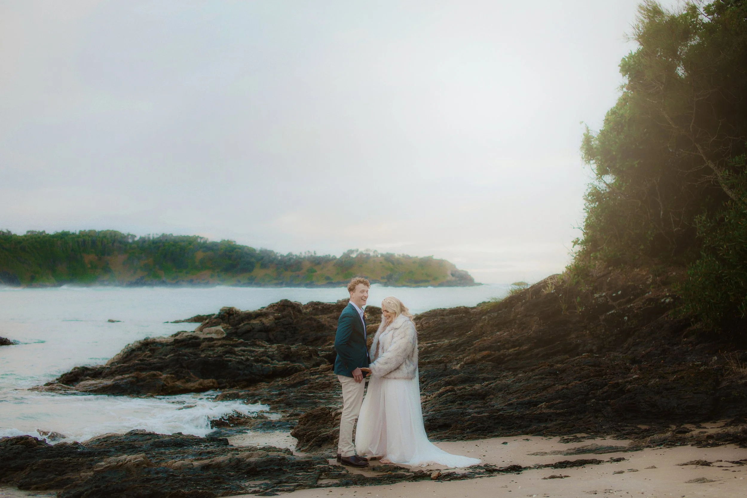 A couple dressed in wedding attire, holding hands, standing on a rocky shoreline near a beach with trees and water in the background.
