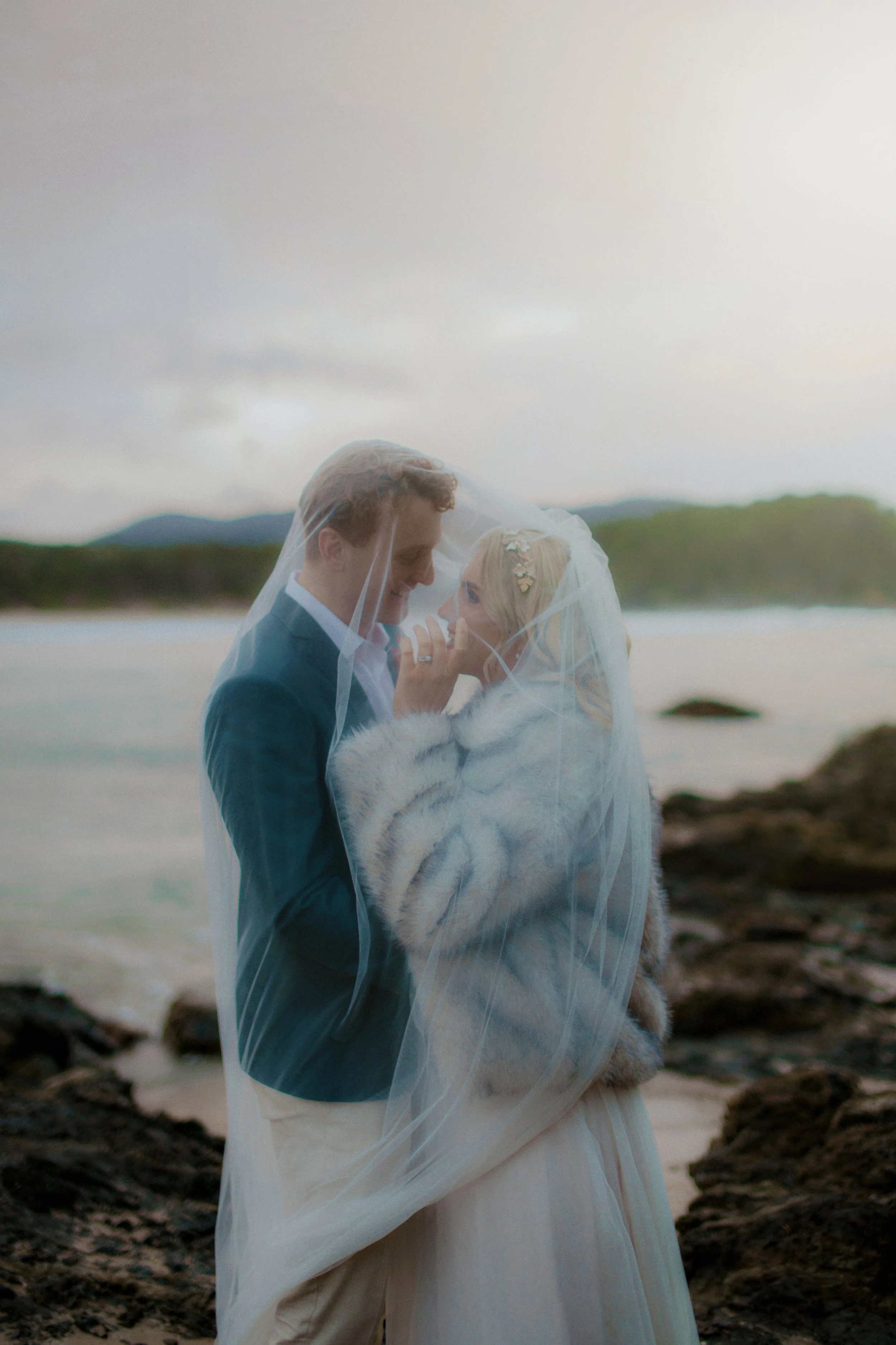 A bride and groom embracing on a beach, with the bride wearing a white wedding gown and faux fur shawl, and the groom in a dark suit, both under a wedding veil.