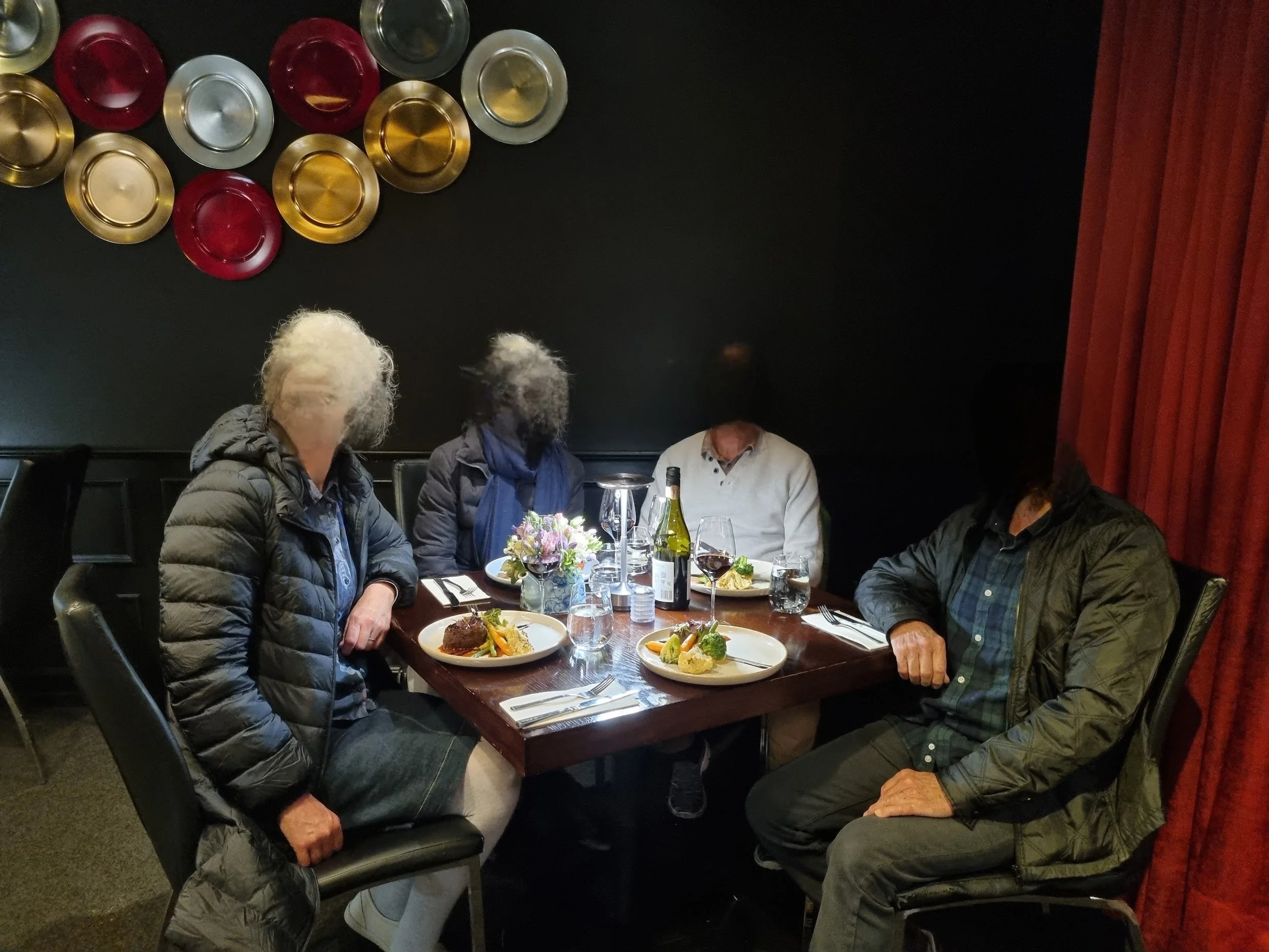 Four people sitting at a dark wood dining table with plates of food, a bottle of wine, and a flower centerpiece, inside a restaurant with black walls and decorative plates.