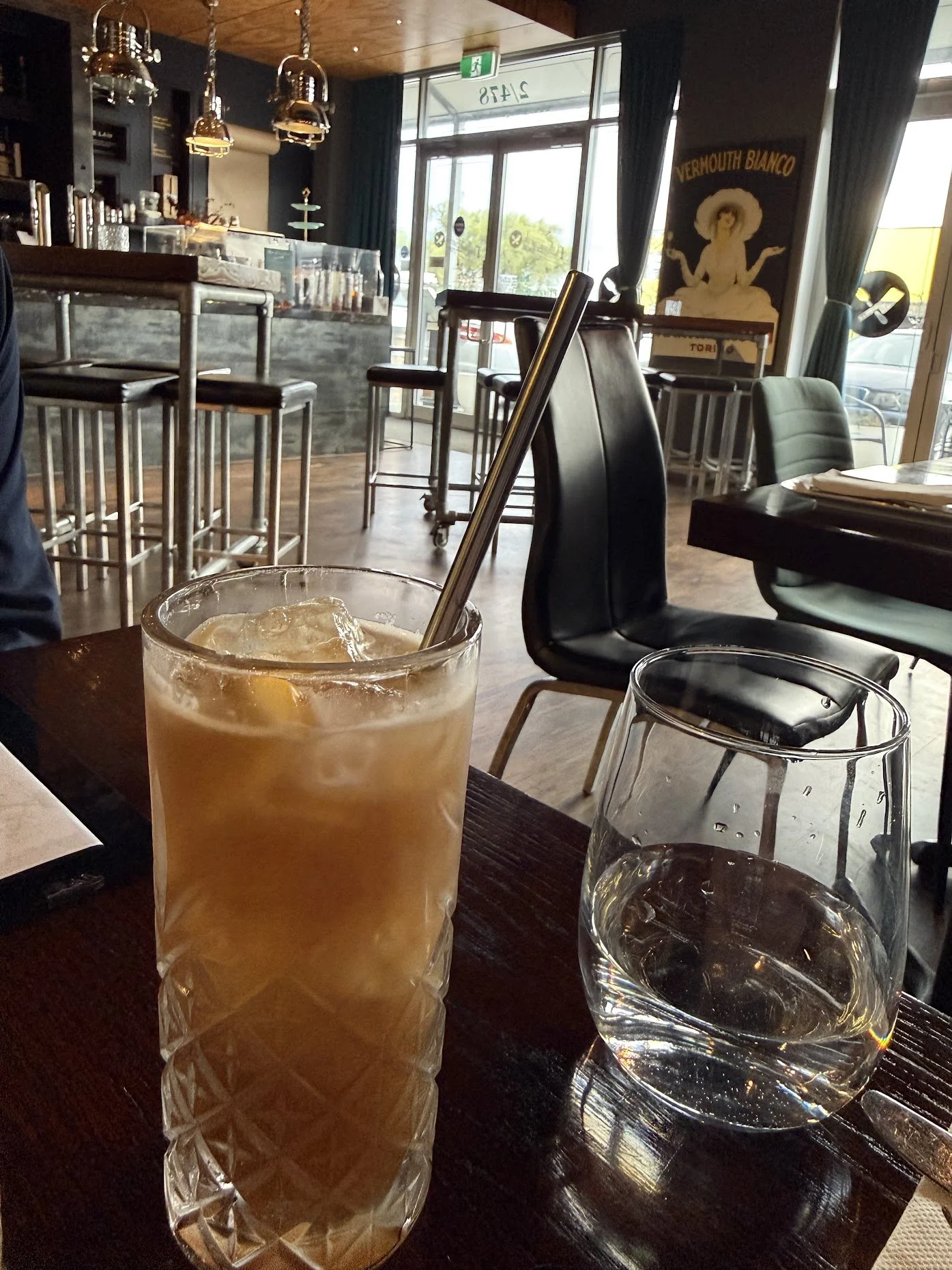 A glass of iced tea with a straw and lemon wedge next to a glass of water on a dark wooden table inside a cafe with natural light coming through large windows.