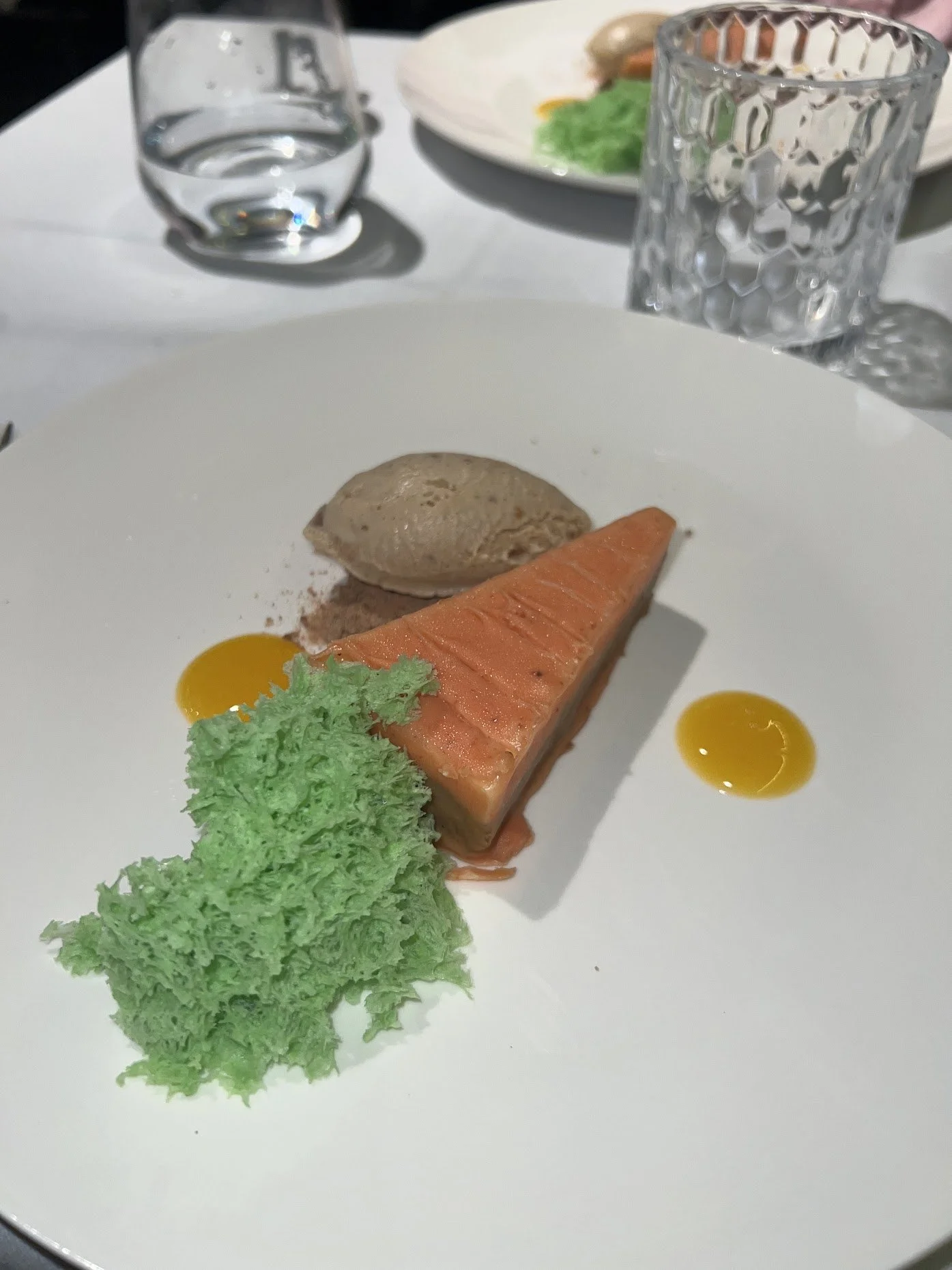 A plate with a slice of salmon, green vegetable, a sphere of sorbet, small yellow sauce dots, and a green garnish, on a white table with glassware and a partially visible dish in the background.