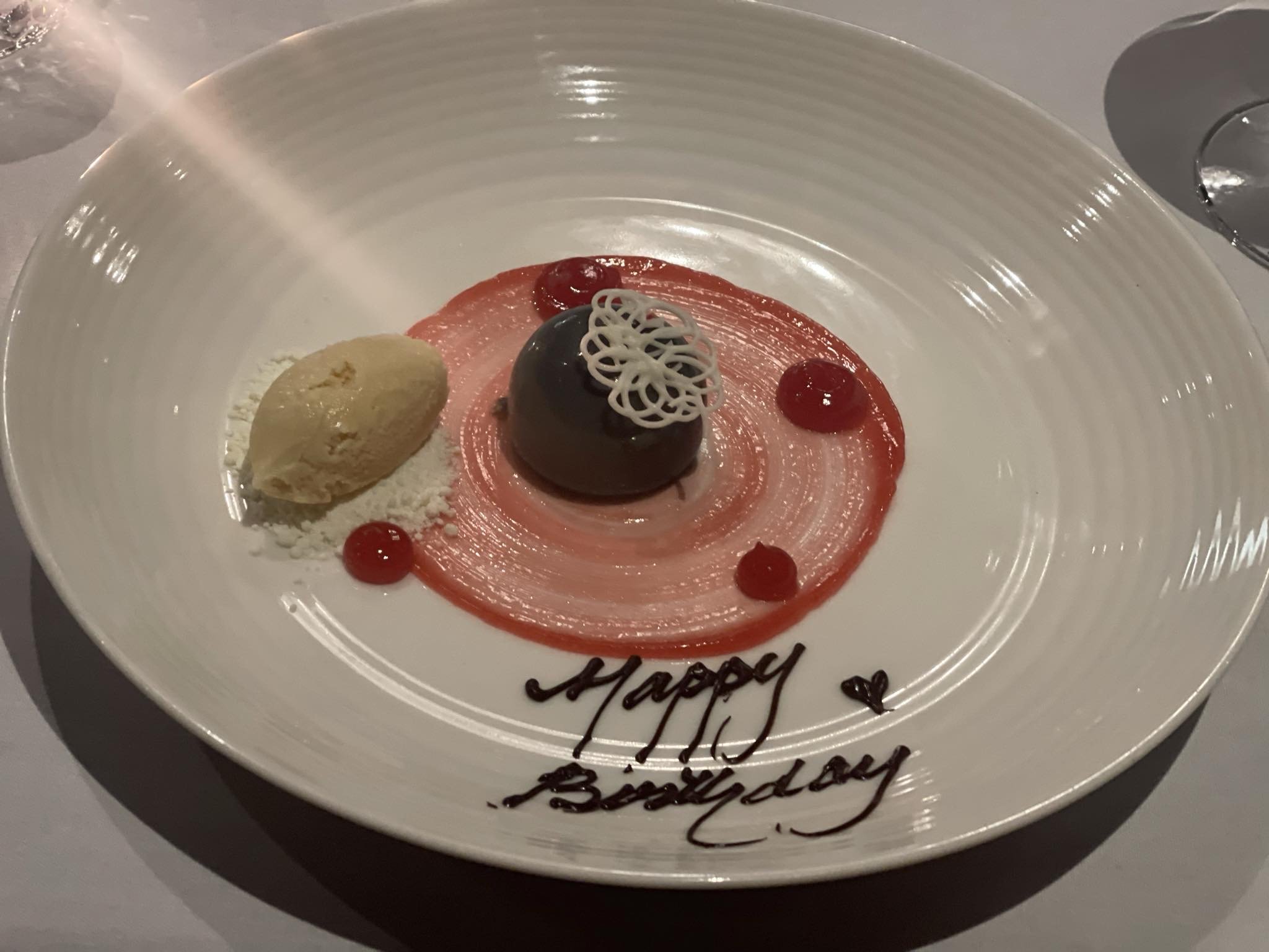 Dessert plate with a scoop of vanilla ice cream, a chocolate dome with white decorative garnish, red sauce drizzled in a circle, whipped cream, and the words 'Happy Birthday' written in chocolate on the plate.