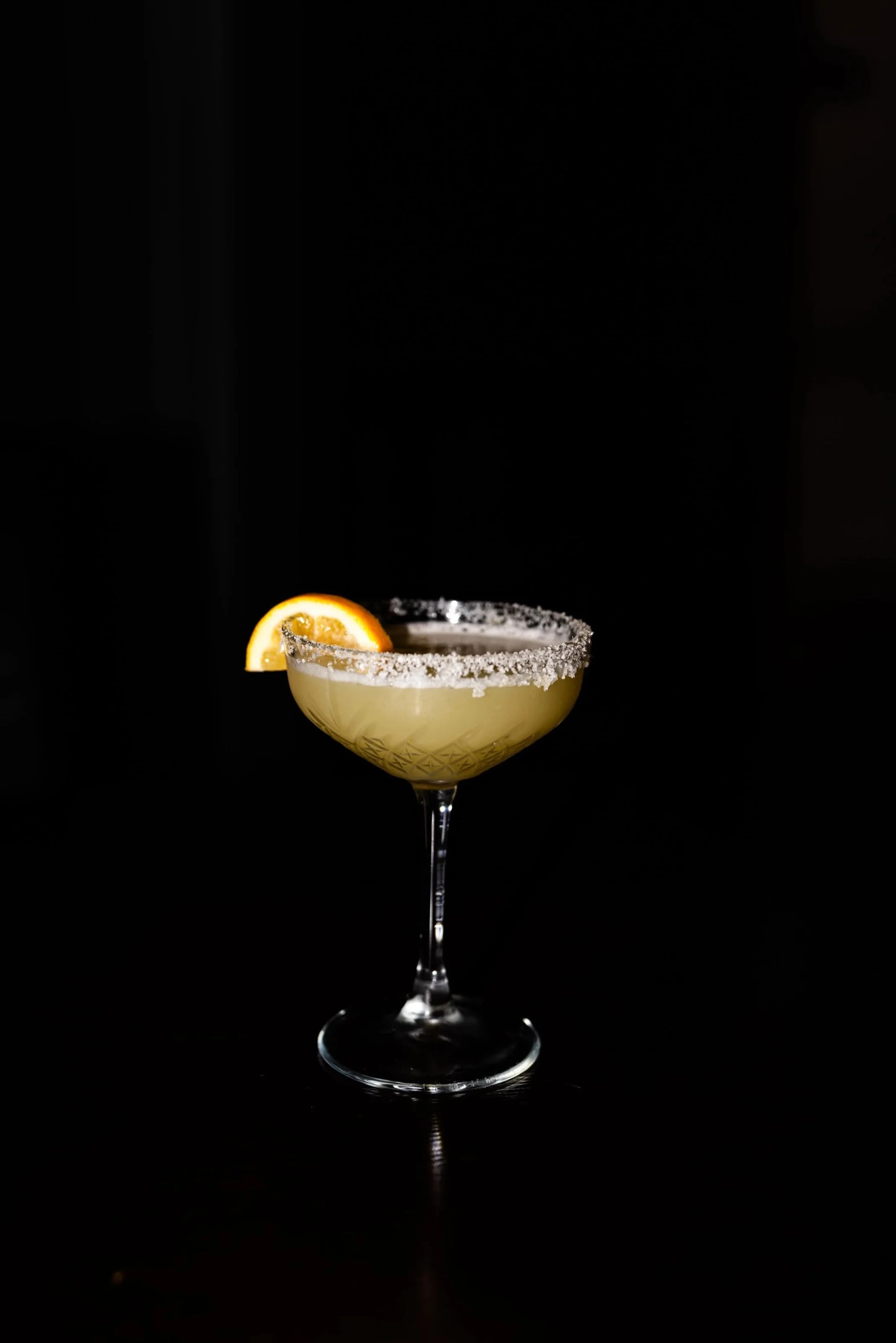 A cocktail in a stemmed glass with a salted rim, garnished with a lemon wedge, set against a black background.