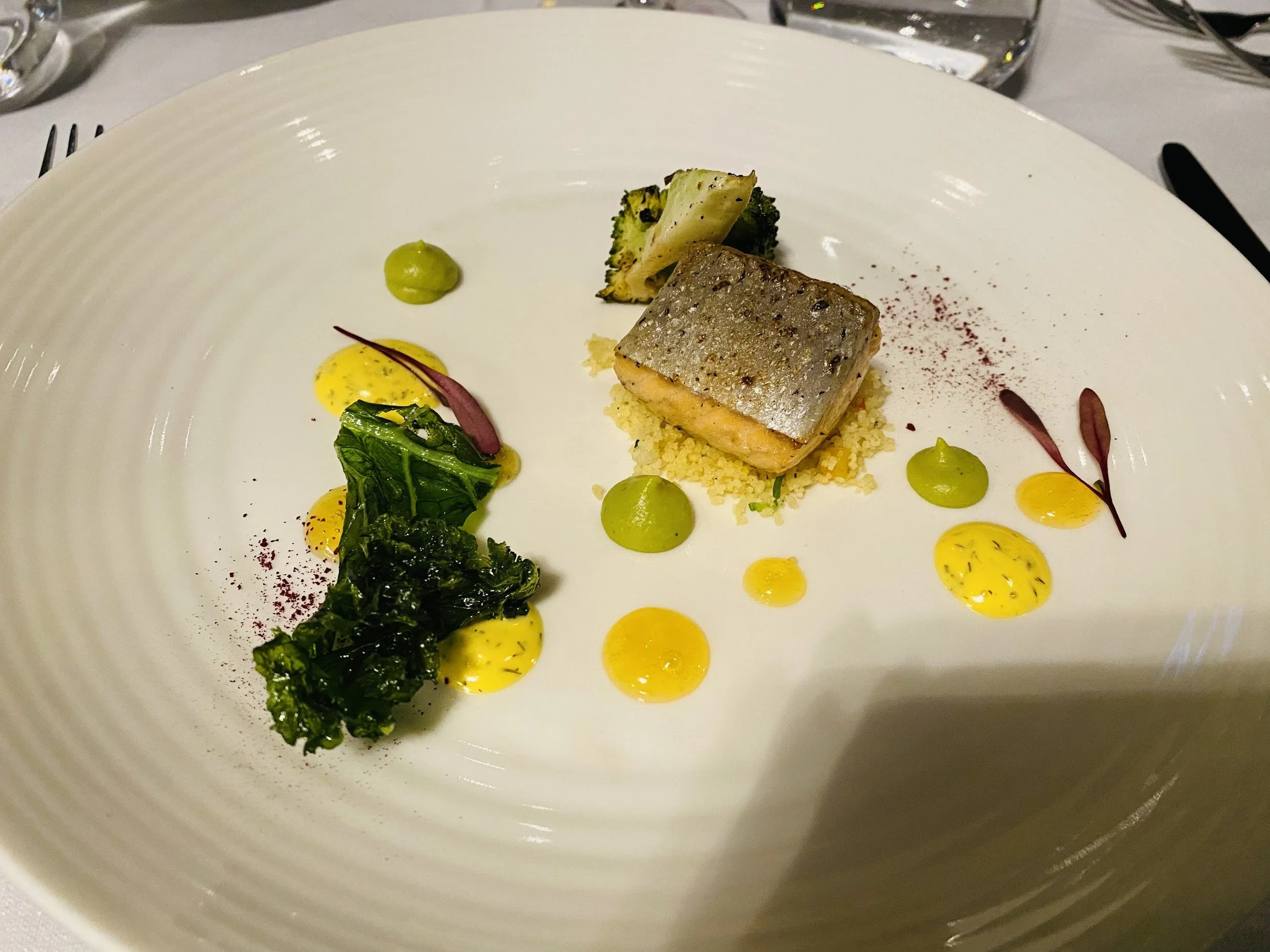 A plated dish featuring a piece of fish with crispy skin, served on a bed of yellow grains, accompanied by assorted vegetables and colorful sauces and garnishes.