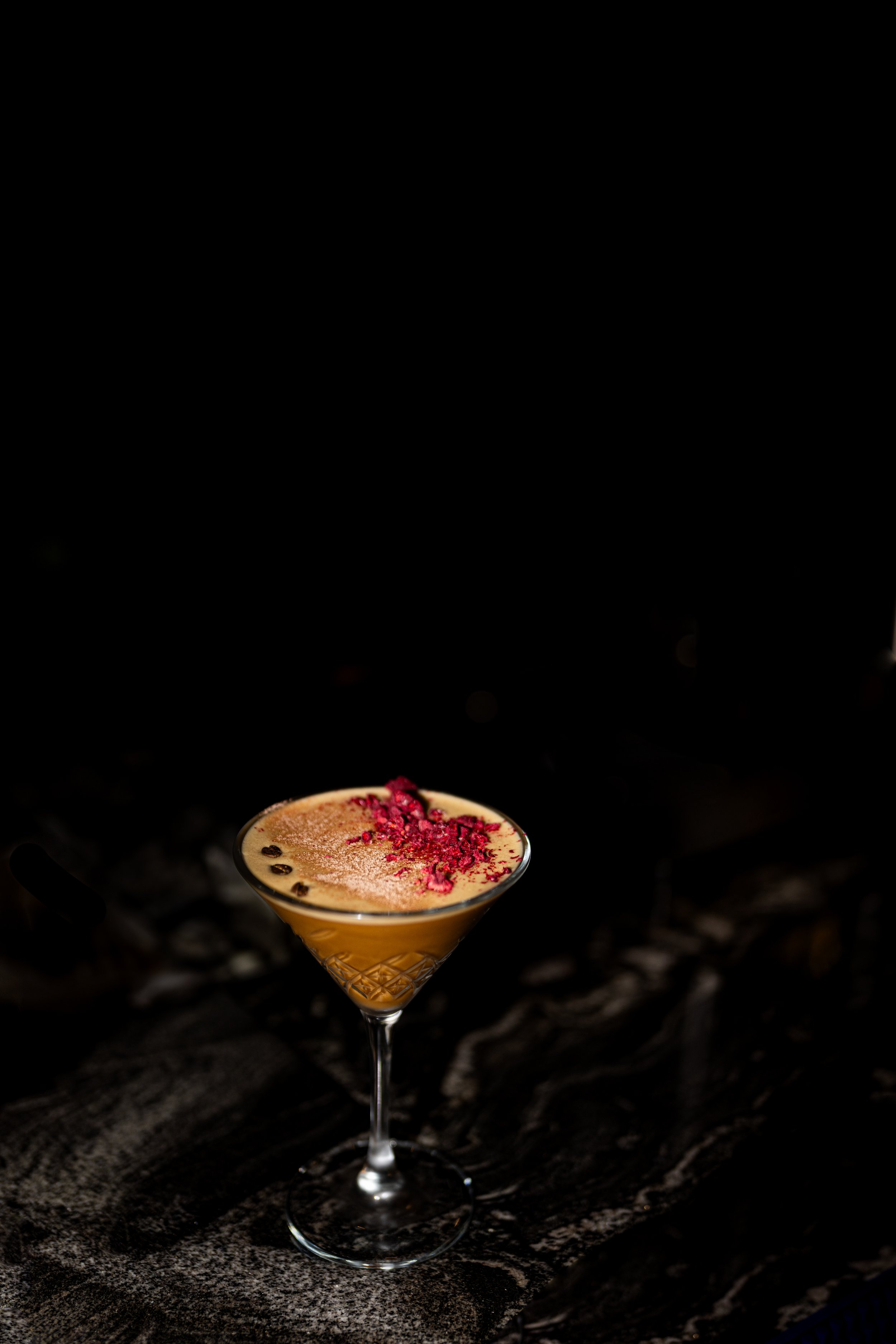 A cocktail glass filled with a light brown drink, garnished with red crumbs and dark seeds on top, sitting on a dark textured surface against a black background.