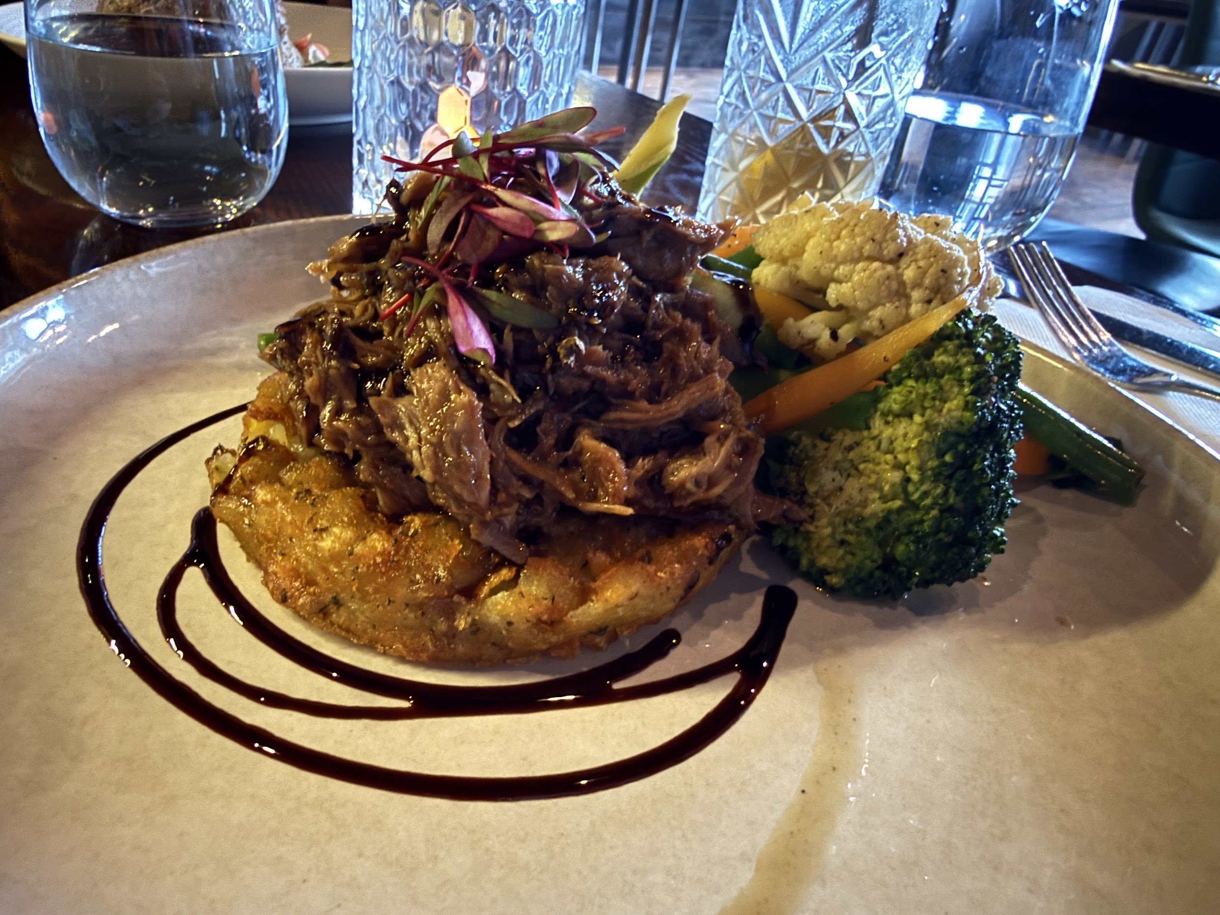 A plated dish with pulled meat on top of a patty, garnished with microgreens, accompanied by broccoli, cauliflower, and carrots, with a drizzle of balsamic glaze on the plate. In the background, glasses of water and a glass with textured pattern are visible.