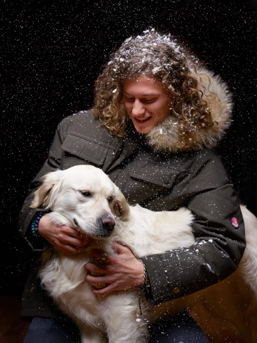 A person with curly hair in a jacket with a fur-lined hood holding a golden retriever dog in a snowy setting.