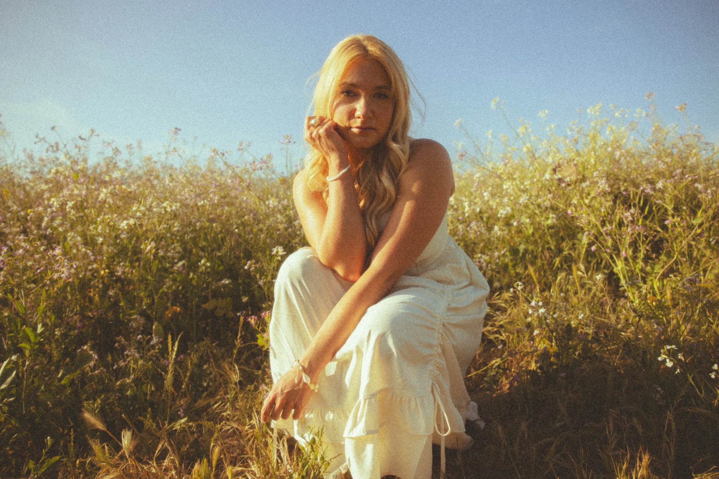 A woman with long blonde hair sitting in a field of flowers on a sunny day.