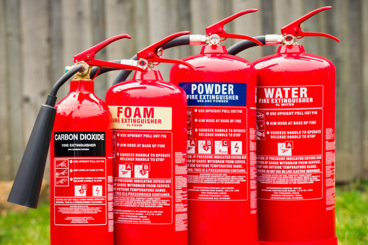 Four red fire extinguishers with different labels and a garden fence in the background.