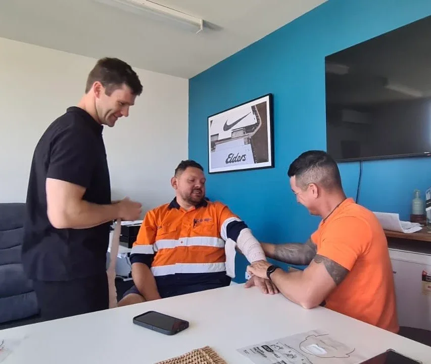 Two men sitting at a table exchanging a handshake or fist bump, with a third man standing nearby. The man on the left is wearing an orange and black work shirt and has a bandaged arm. The man on the right is wearing orange T-shirt with tattoos and is checking the other man's bandaged arm. They are in a room with a blue wall, a framed Nike logo, and a large TV.