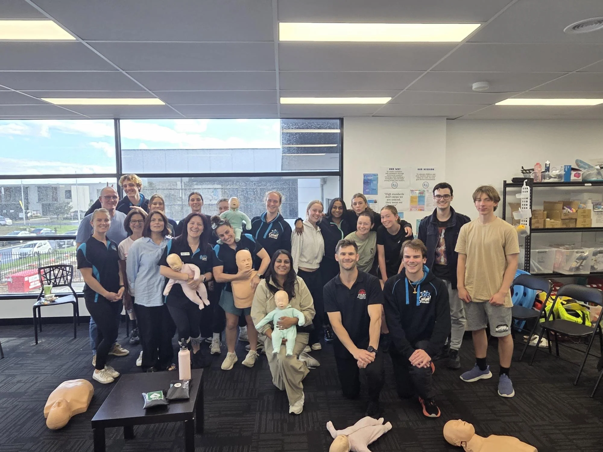 Group of people in a classroom or training room posing for a photo with CPR mannequins and training supplies on the table in front of them.