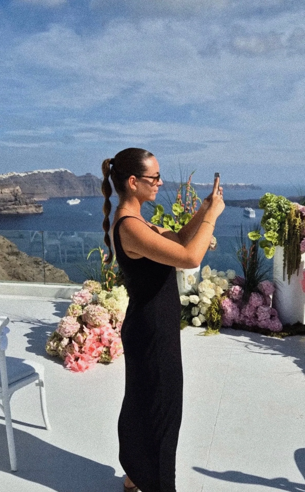 A woman in a black dress taking a photo with her phone at a seaside terrace with floral arrangements, overlooking the ocean and cliffs.
