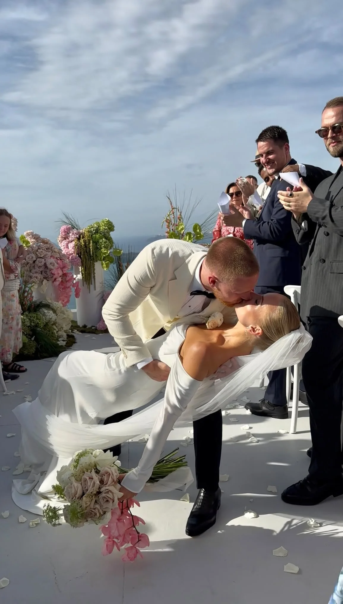 A bride and groom kiss during their wedding ceremony outdoors, with guests in suits and dresses clapping and smiling, floral arrangements, and a sky with clouds in the background.