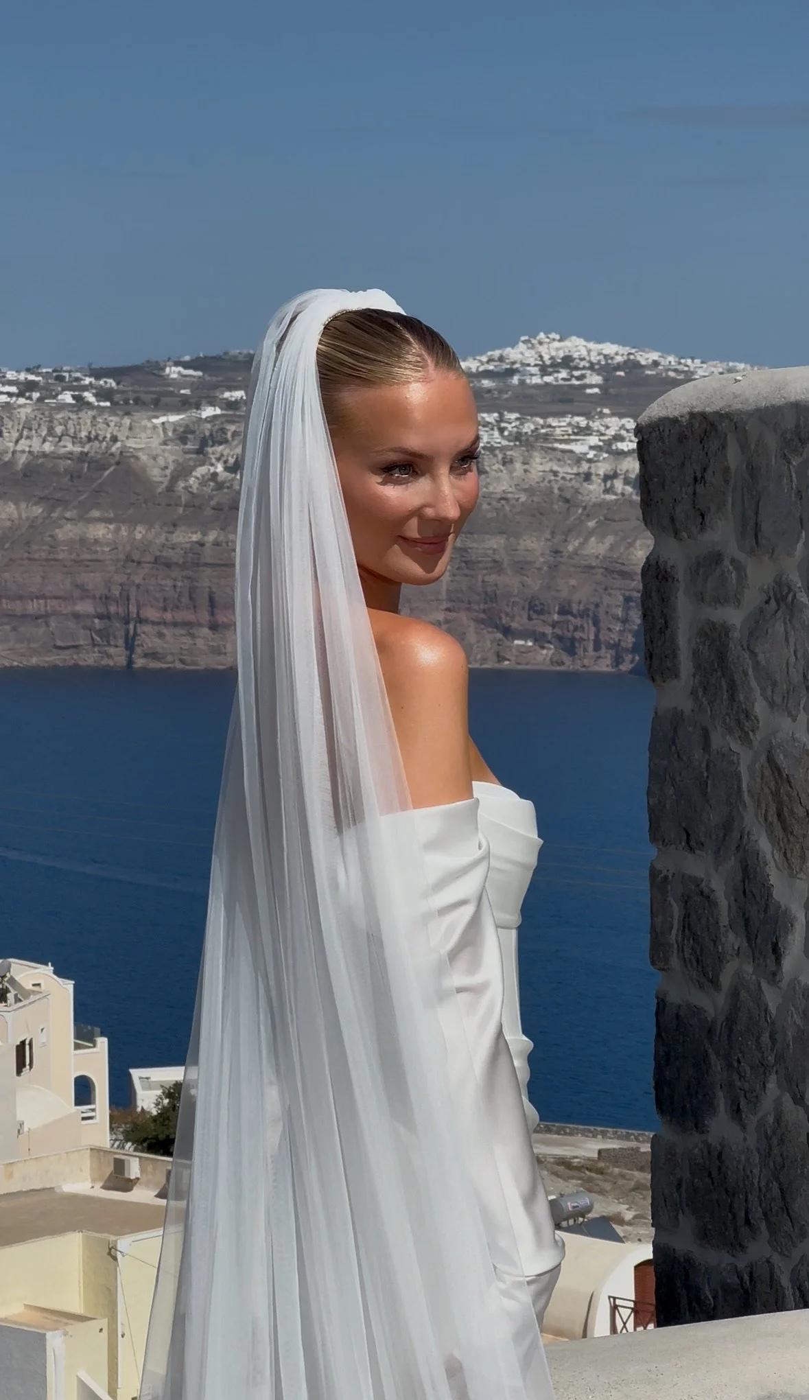 A bride wearing a white dress and long veil standing near a stone wall with a scenic ocean and island background.