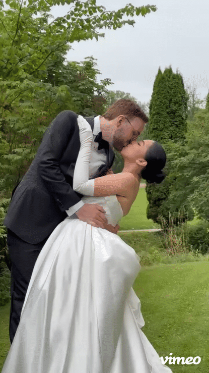 A newlywed couple sharing a kiss outdoors on a grassy area with trees in the background. The groom is in a tuxedo, and the bride is in a white wedding gown.
