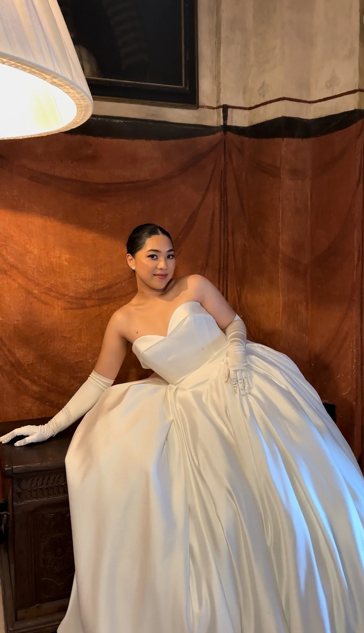A woman sitting on a wooden cabinet, wearing a white satin strapless ball gown, long white gloves, and earrings, with a warm brown textured wall behind her and a large lamp partially visible in the top left corner.