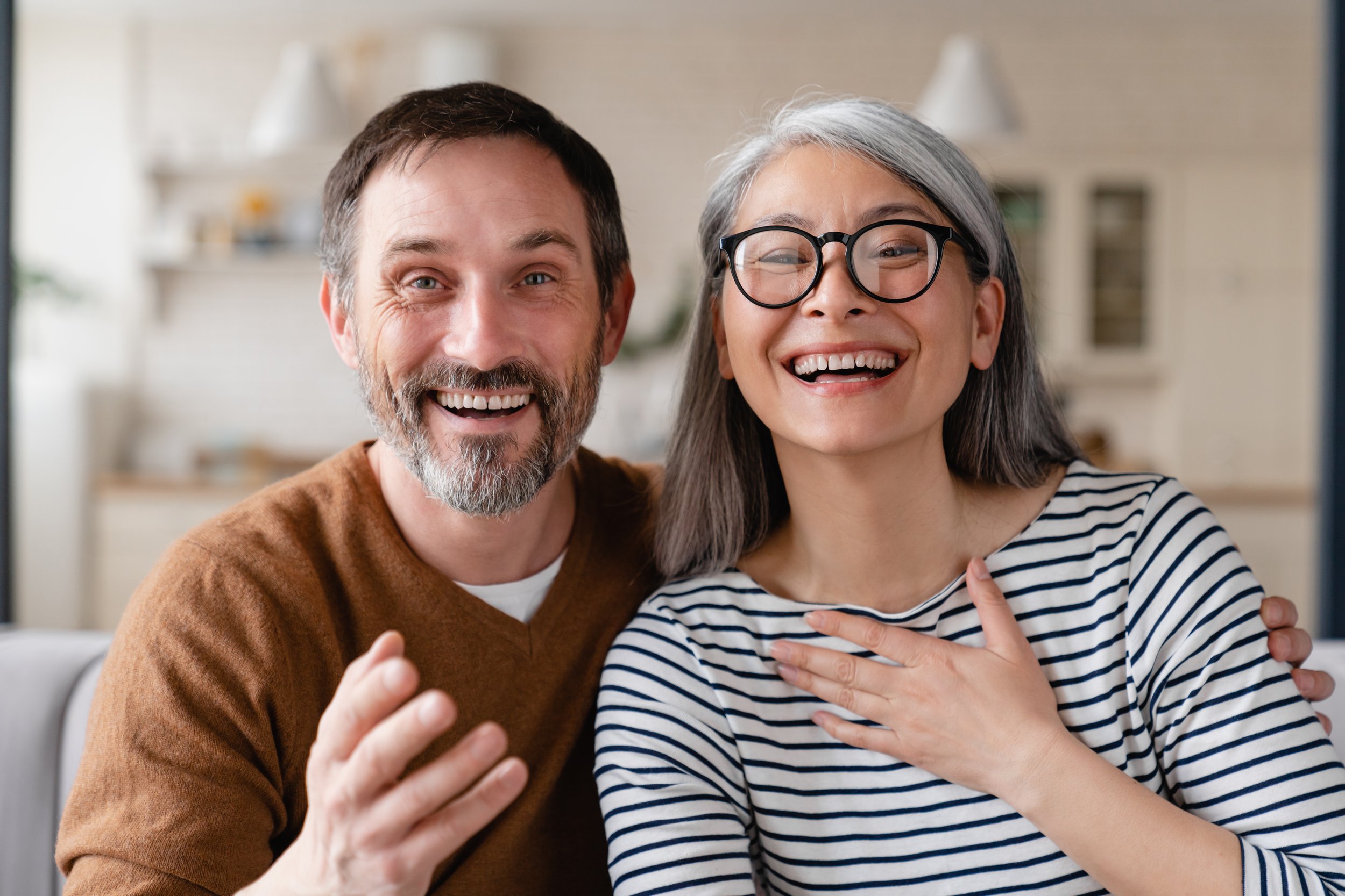A smiling man and woman looking at a camera, with the woman touching her chest and the man making a gesture with his hand in a cozy indoor setting.